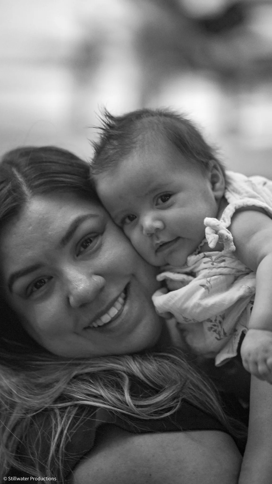 A woman and a baby lying close together, smiling and looking at the camera in a black-and-white photo.