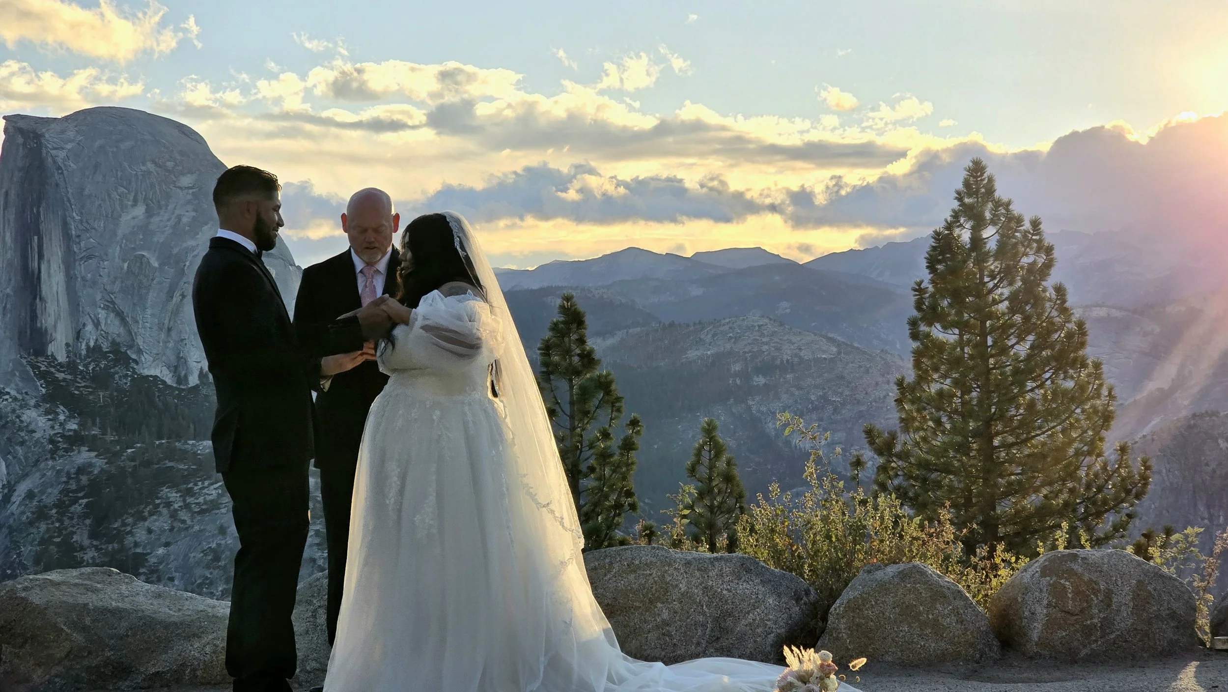 A couple getting married outdoors as the sun sets behind mountains. The bride is in a white gown with a veil, and the groom is in a black suit. An officiant stands between them, holding hands. Trees and rocks are in the foreground with a scenic mount