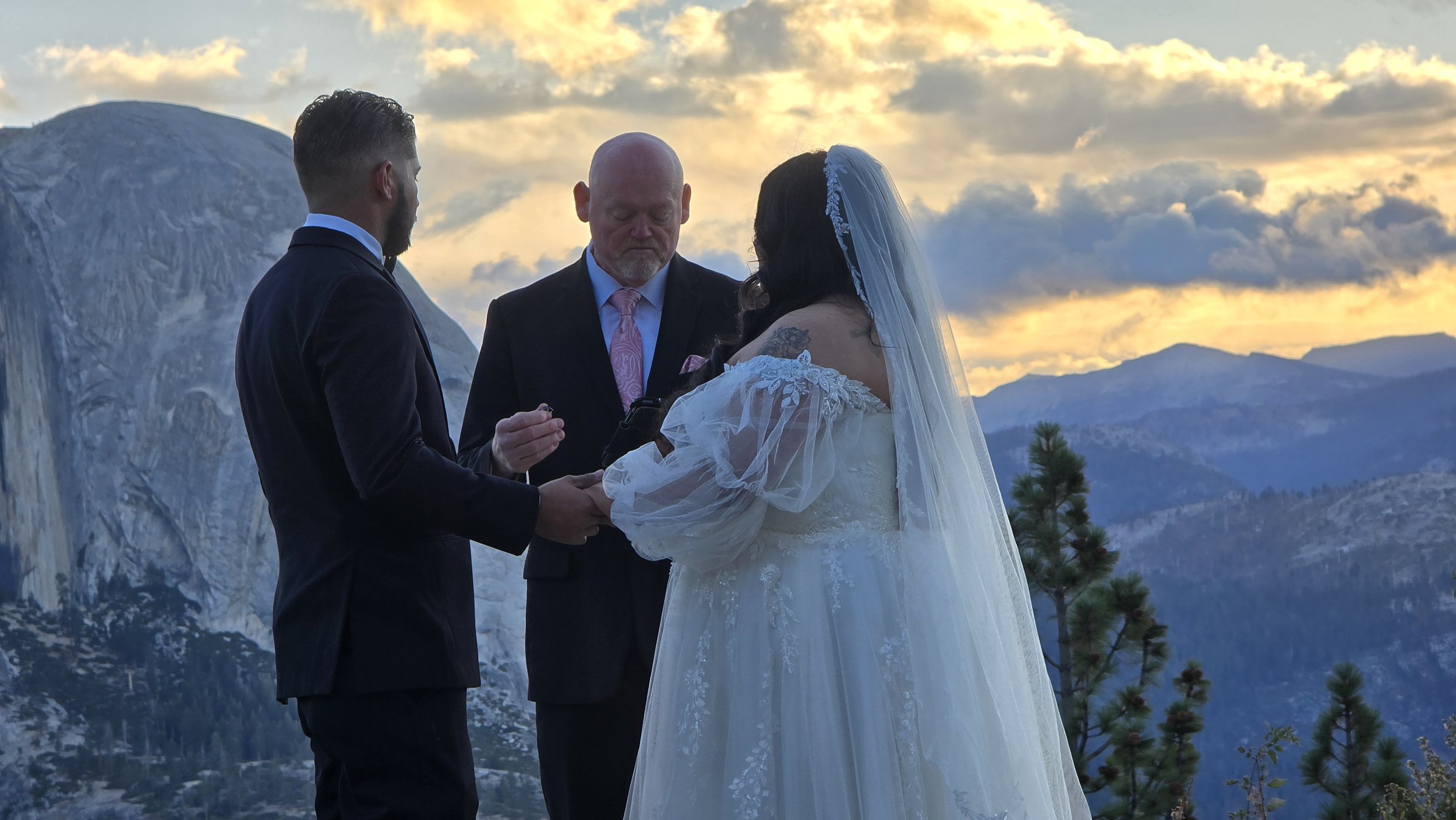 A couple getting married outdoors at sunset, with mountains in the background. The groom is in a dark suit and the bride is in a white wedding dress with lace and a veil, holding hands and facing each other.