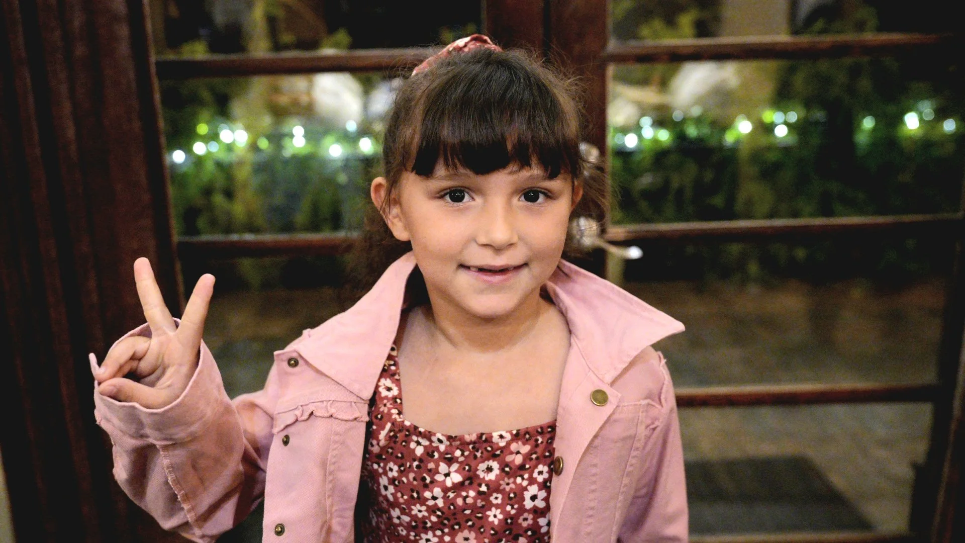 A young girl with brown hair and bangs smiling and making a peace sign with her right hand. She is wearing a light pink jacket over a red and white floral dress, and is standing indoors near a wooden door with glass panes. The background shows blurry