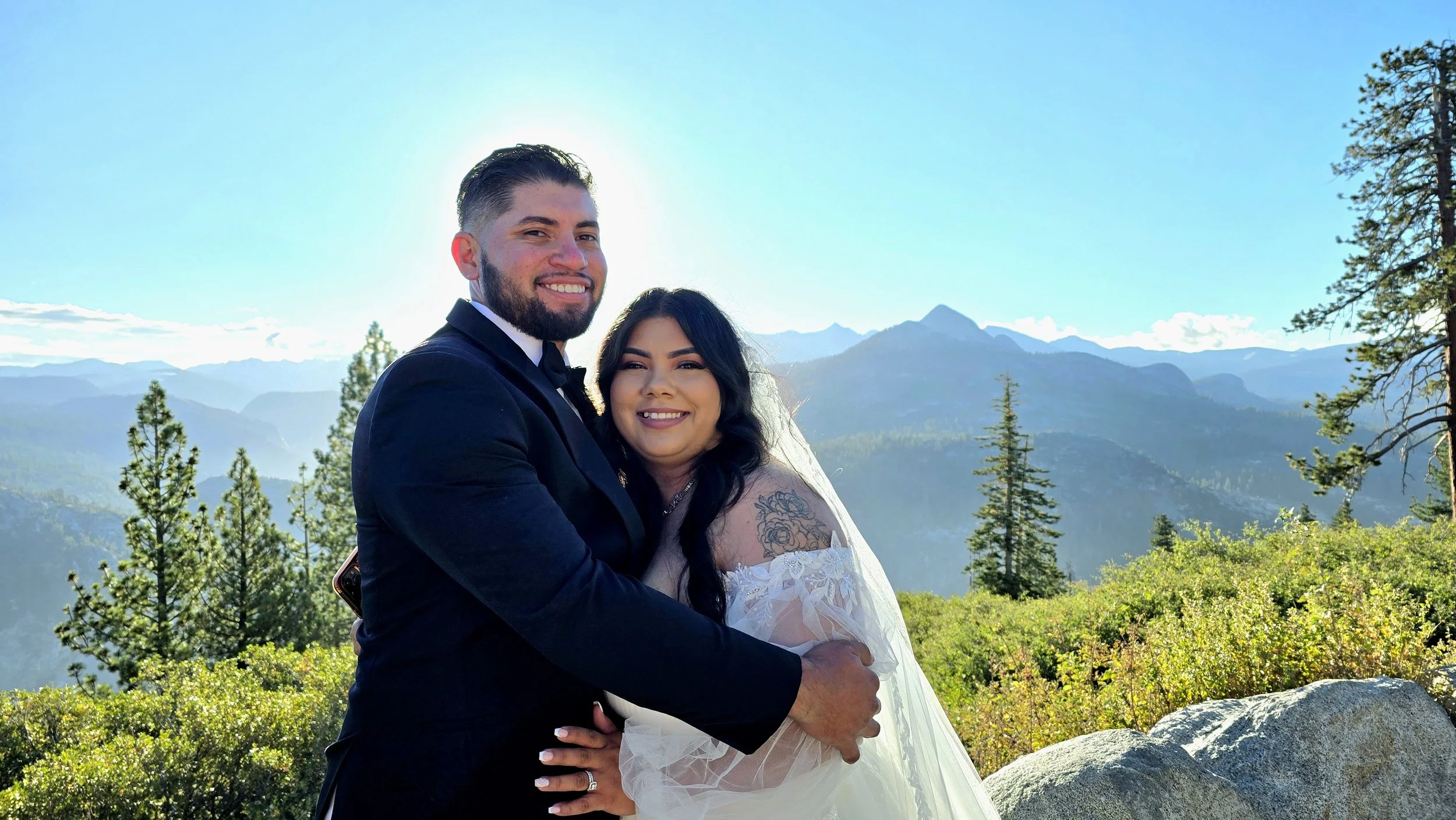 A smiling couple in wedding attire hugging outdoors against a scenic mountain landscape with blue sky, trees, and rocky terrain.