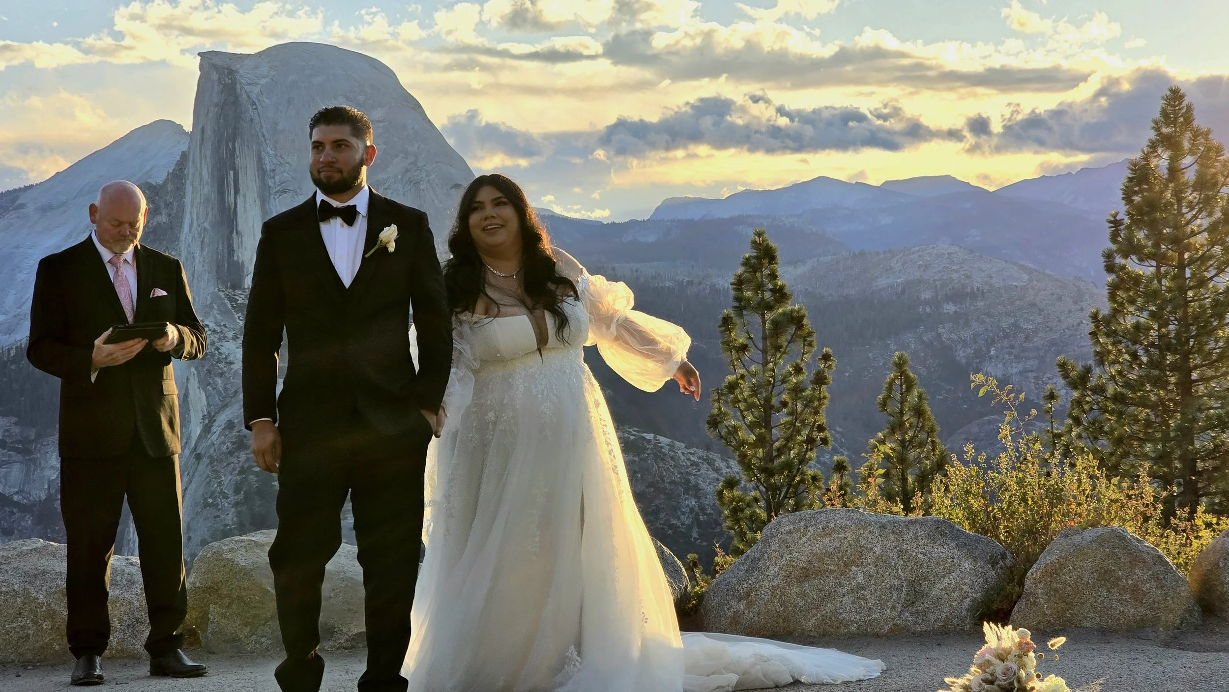 A wedding ceremony outdoors in front of a mountain, with a bride and groom standing together. The groom wears a black tuxedo with a white shirt and black bow tie, and the bride wears a white wedding dress with puffy sleeves. An officiant in a black s