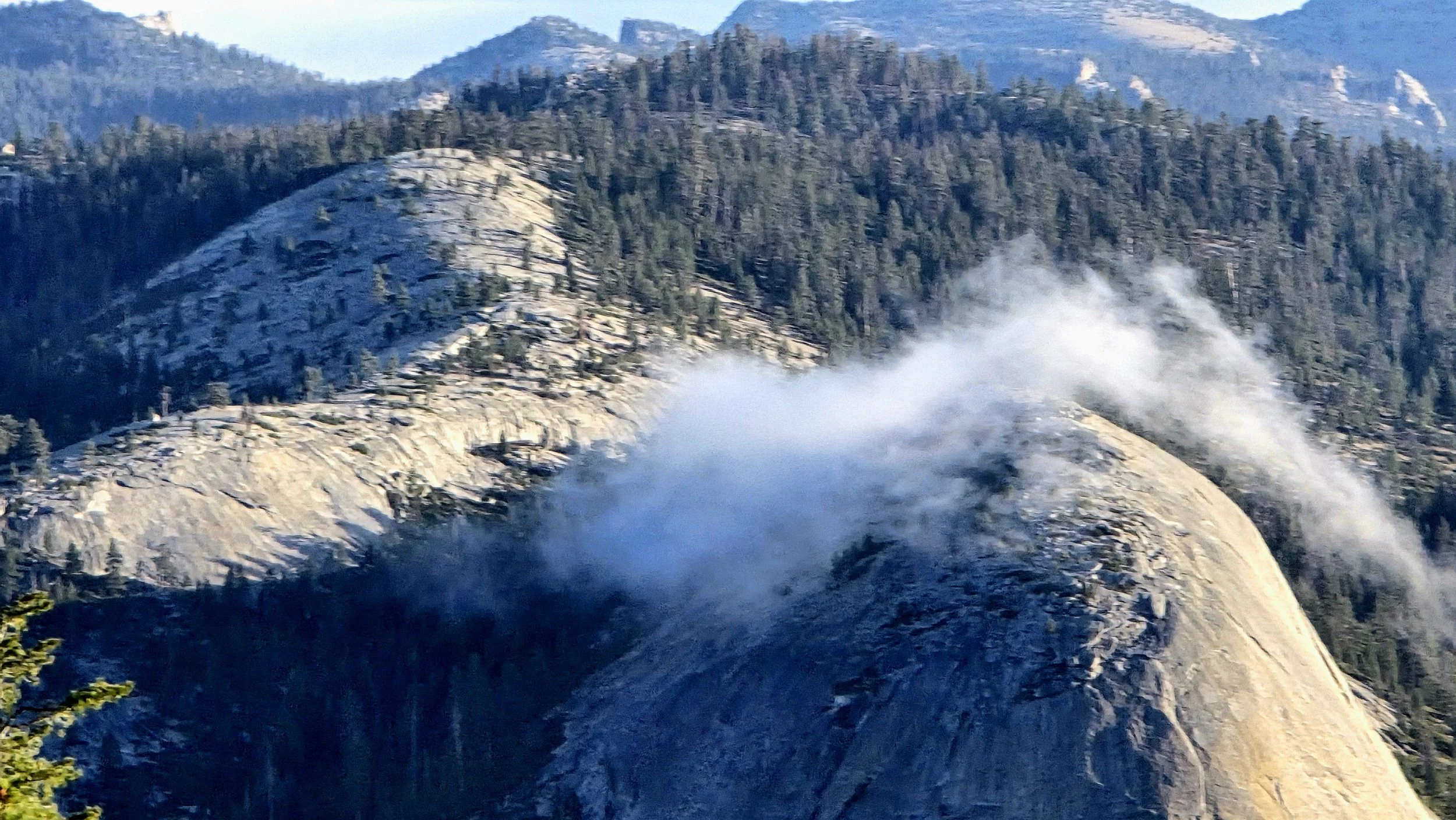 A mountainous landscape with trees and rugged rock formations, with clouds of steam or smoke rising from part of the mountain.