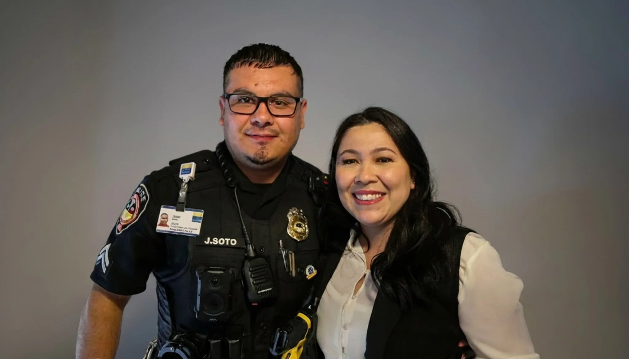 A man in a police uniform and a woman in a light-colored shirt pose together and smile in front of a plain gray background.