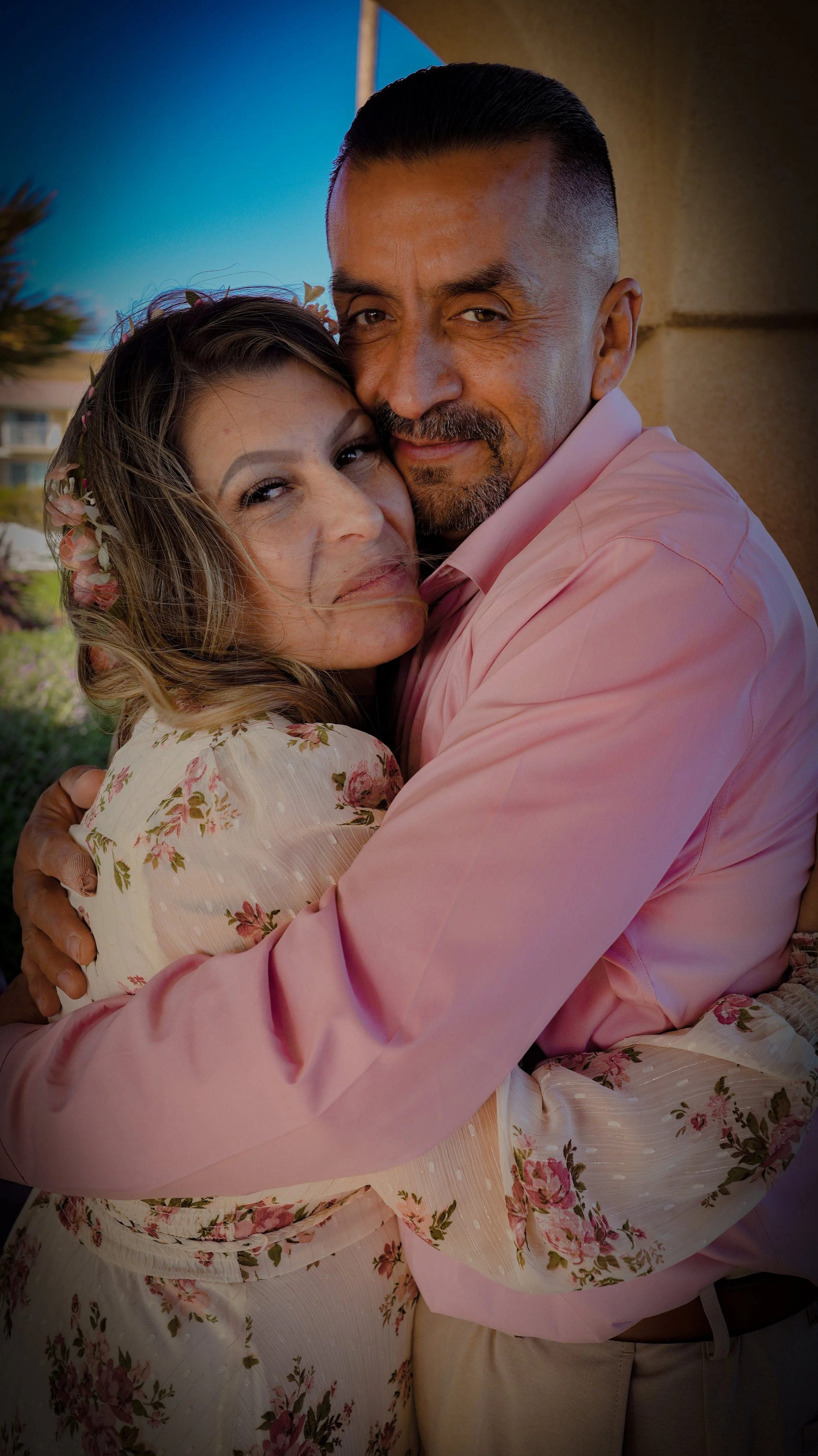 A man and woman hugging each other closely outside, smiling warmly at the camera. The woman has long, wavy hair with flowers and is wearing a floral dress. The man has short dark hair, a goatee, and is wearing a pink shirt. The background shows a bui