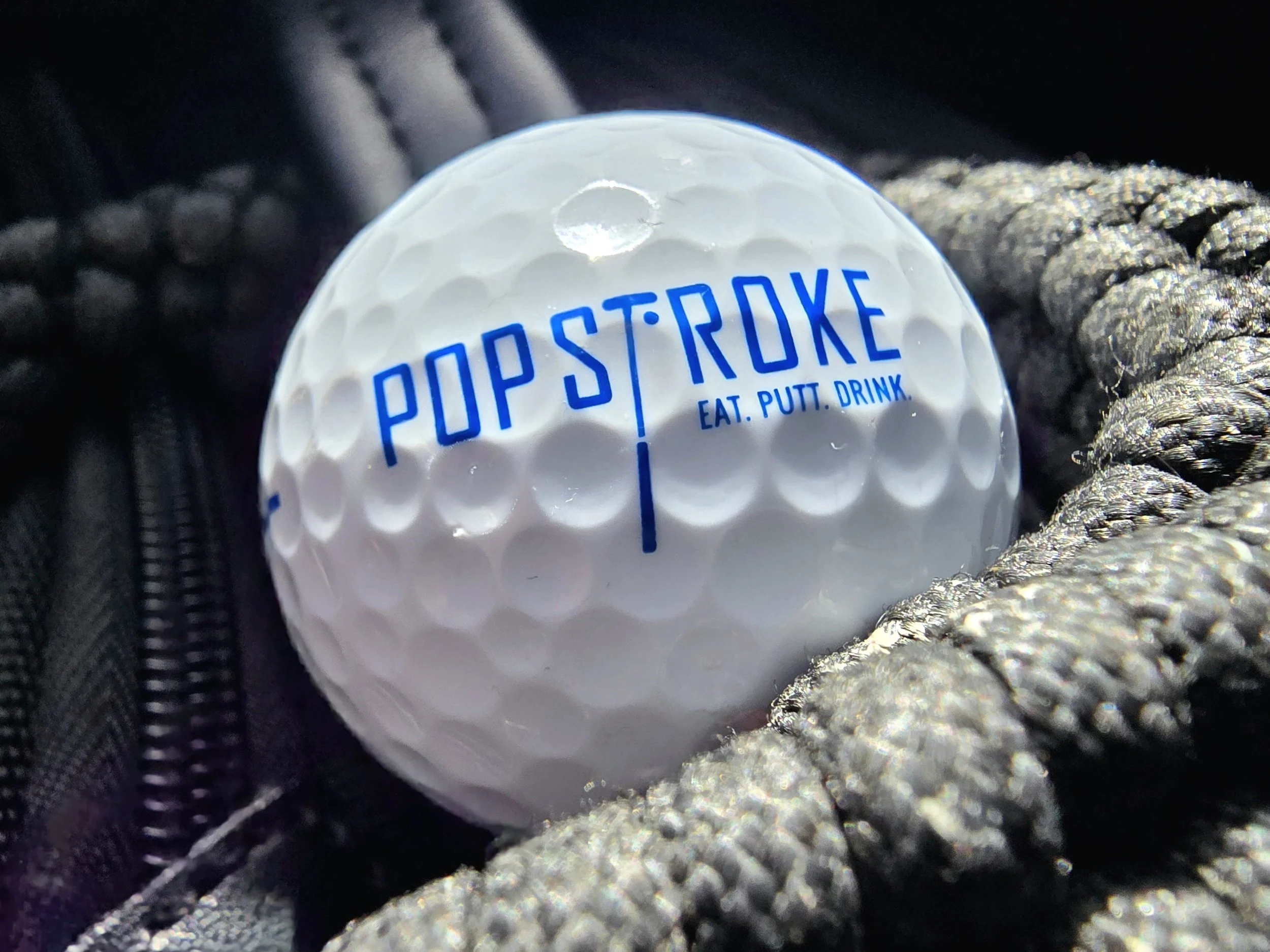 Close-up of a golf ball with the text "POP STI ROKE" and the words "EAT. PUTT. DRINK." printed in blue, resting on a textured surface with dark fabric and metallic braided rope nearby.