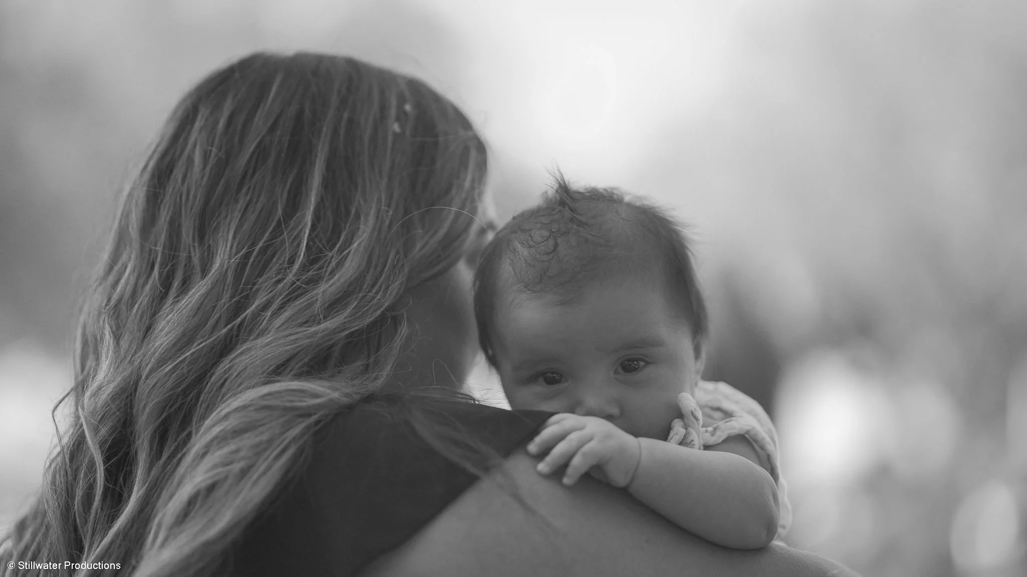 A woman with long hair holds a young child in her arms, cradling the child's face close to her. The child has curly hair and is looking directly at the camera. The background is blurred, emphasizing the subjects.