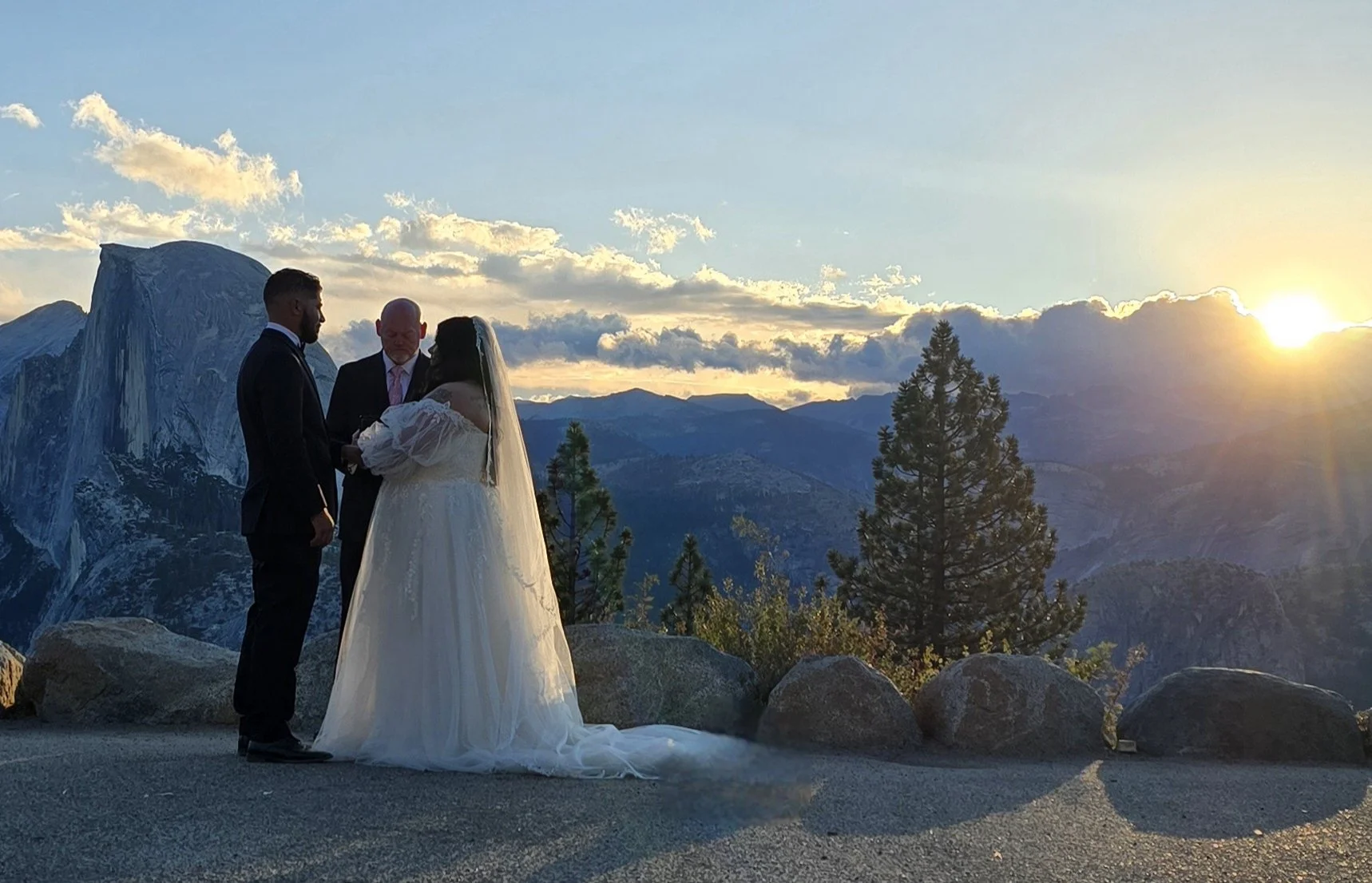 A couple in wedding attire standing outdoors at sunset, with a mountain range and trees in the background.
