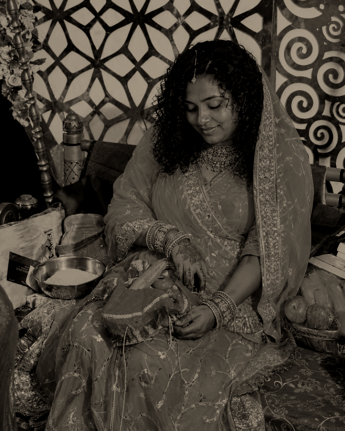 Een vrouw in traditionele Indiase kleding zit op een bed, omringd door fruit, lotusbollen en andere items, met een decoratieve achtergrond.