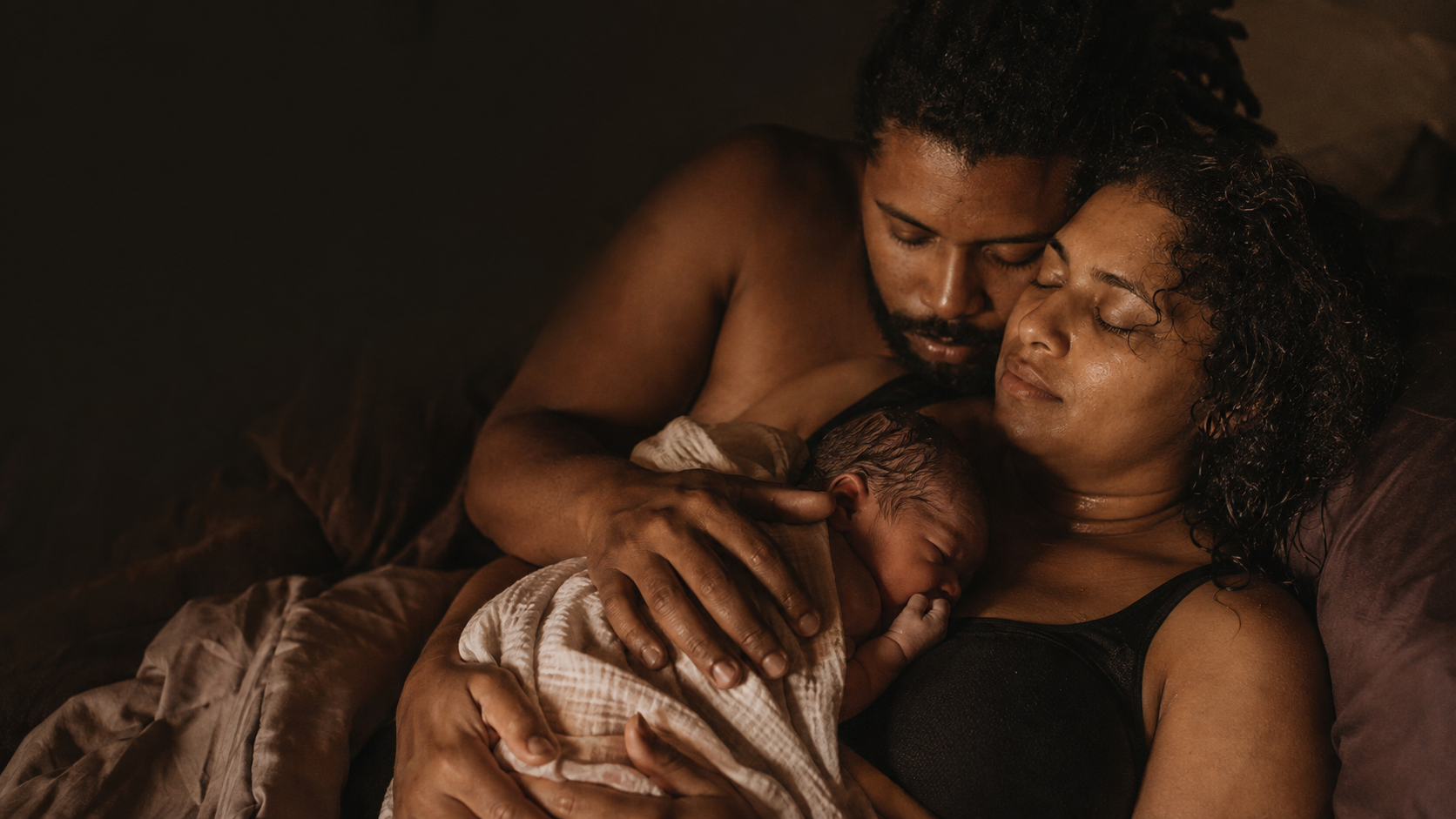 Een pasgeboren baby wordt met liefde door haar ouders vastgehouden en gekoesterd, beiden met gesloten ogen en een gevoel van vrede.
