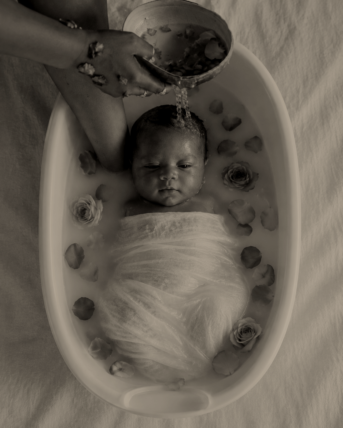 Baby wordt in een bad met bloemen en rozenblaadjes gewassen, terwijl een volwassene water over het hoofd giet.