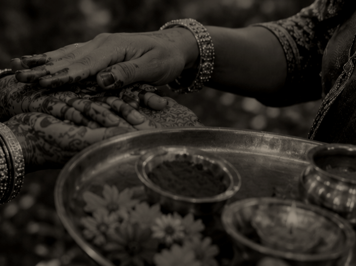 Twee handen met traditionele sieraden en henna-tatoeages die elkaar aanraken, naast een dienblad met verschillende kleine bakjes met kruiden en bloemen, waarschijnlijk tijdens een traditioneel Indiase of Hindoestaanse ceremonie.
