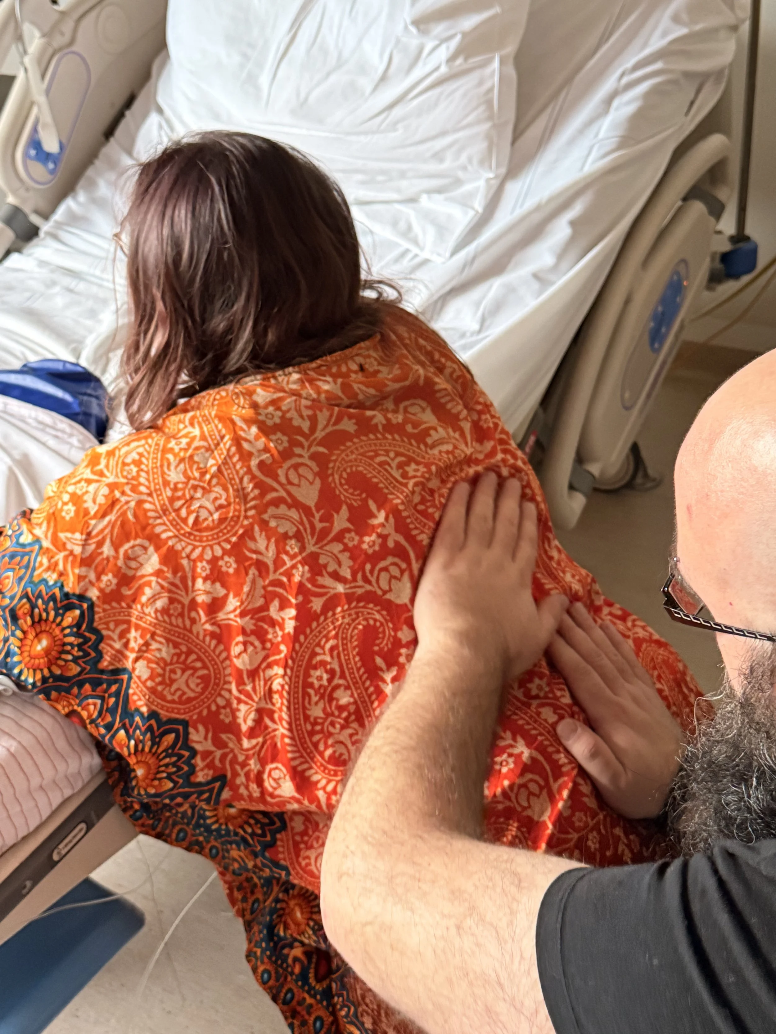 Persoon met grijs haar en een smartwatch geeft een knuffel aan een vrouw die op een ziekenhuisbed ligt, gekleed in een oranje-gekleurde shawl met een patroon.