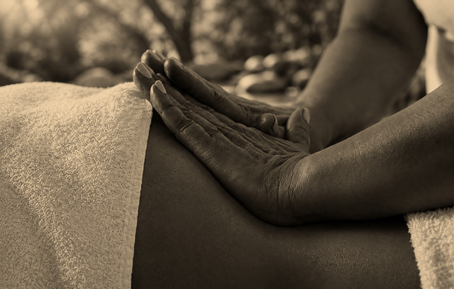 Close-up van twee handen die een massage uitvoeren op een rug, met een handpak en een achtergrond van bomen, in sepia-tonen.