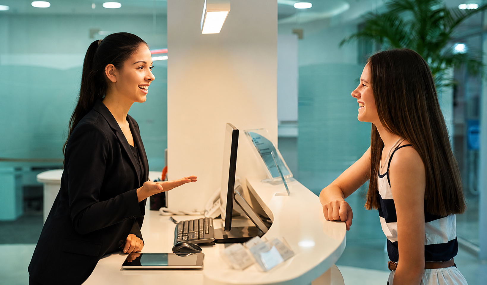 A young woman with long, dark hair and a striped sleeveless top is smiling and talking to a smiling female receptionist at a modern reception desk in an office or hotel lobby.