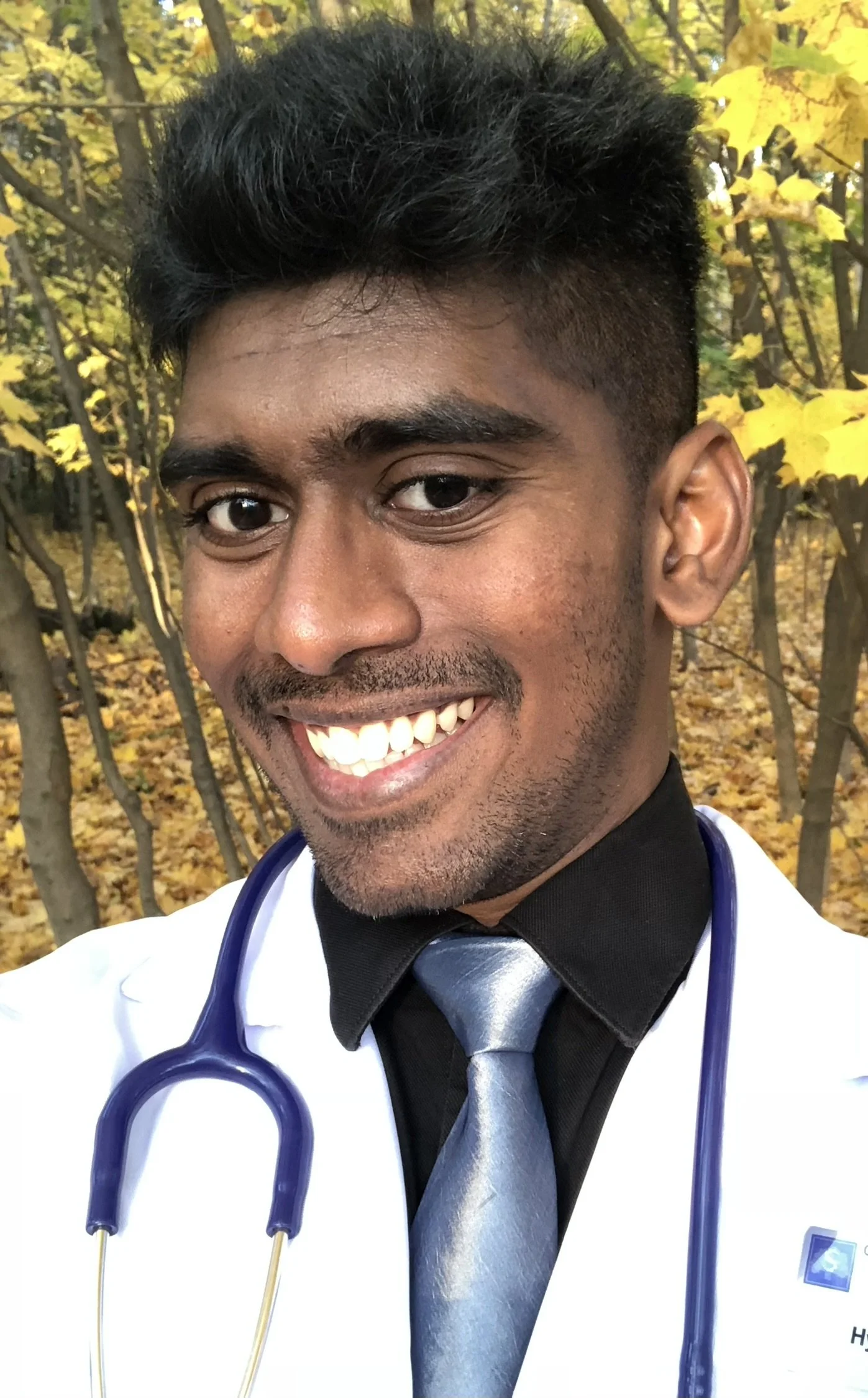 A smiling young man with dark hair, wearing a doctor's white coat, black dress shirt, gray tie, and a stethoscope around his neck. The background shows autumn trees with yellow leaves.