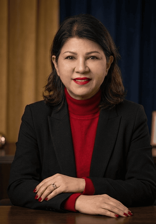 A woman with shoulder-length dark hair wearing a red turtleneck sweater and black blazer, sitting at a wooden table with her hands folded in front of her, smiling against a background of a wood-paneled wall.
Datin Norsiah, Group General Manager