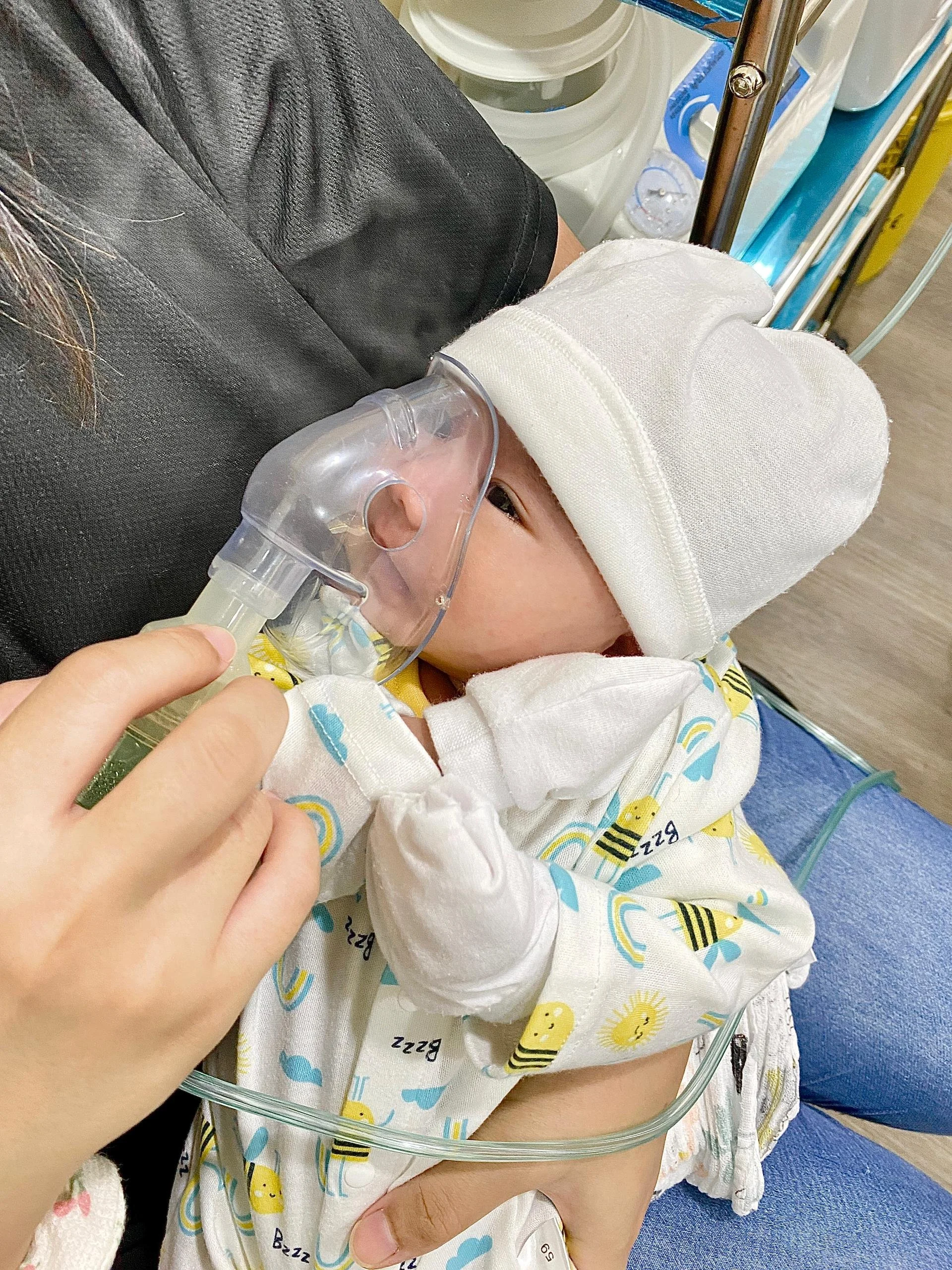 A baby in a hospital bed with an oxygen mask, wearing a white hat and a colorful gown, being held by an adult in a black shirt.