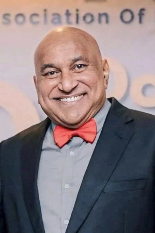 A smiling, bald man wearing a dark suit, light gray shirt, and red bow tie at an event with a backdrop that reads 'sociation of'.