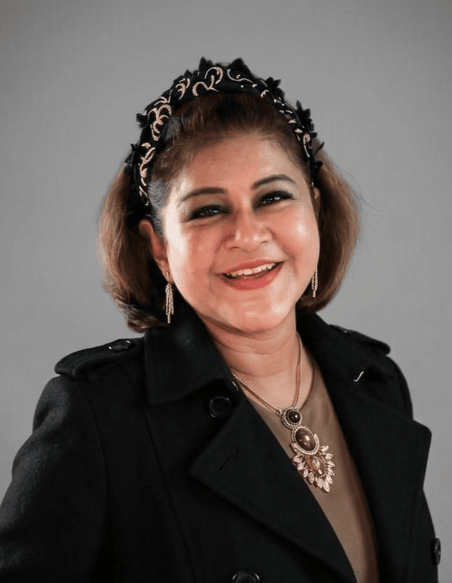 Smiling woman with short brown hair wearing a black coat, a decorative headband, and silver jewelry, against a gray background.