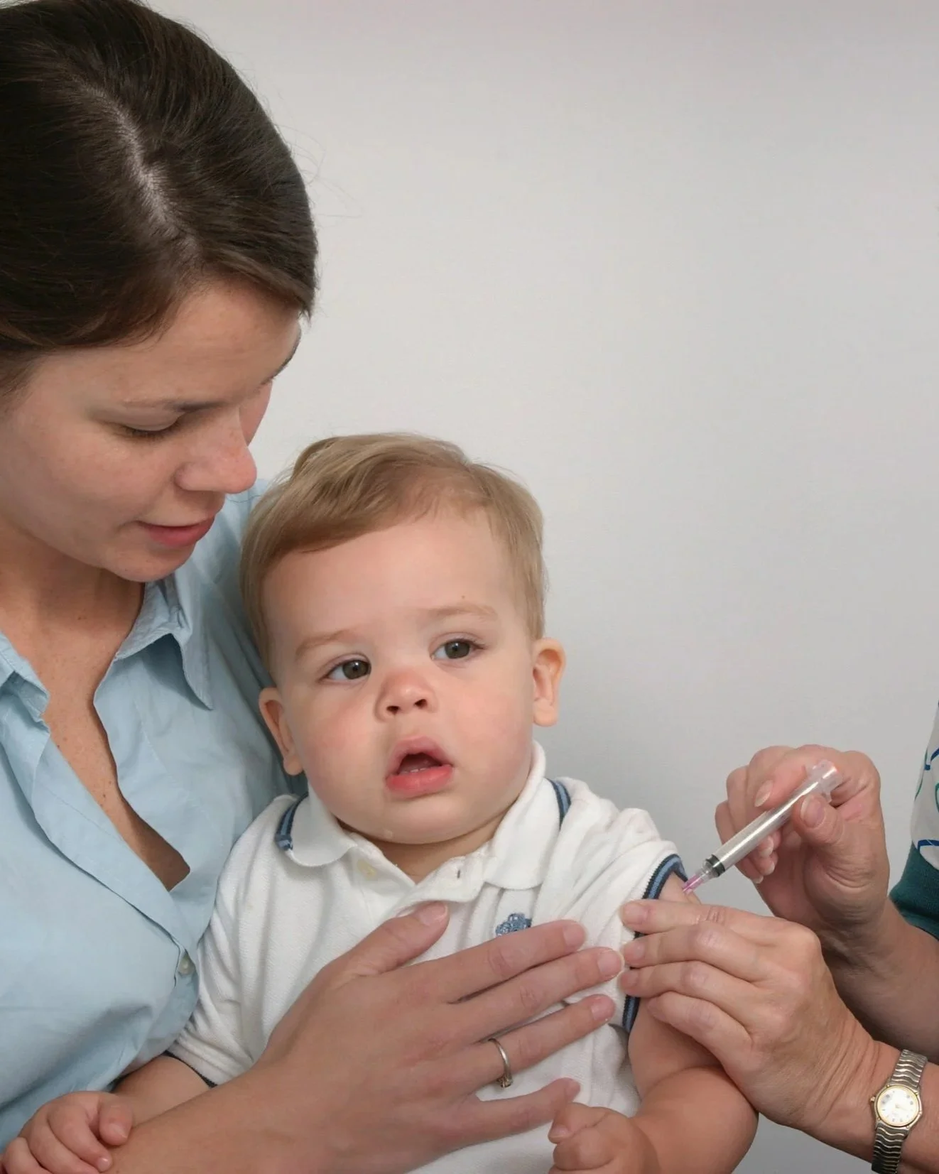 A young boy is being vaccinated by a healthcare professional, while a woman, likely his mother, holds him. The woman is looking at the boy, who has a slightly apprehensive expression. The boy is wearing a white polo shirt with navy accents, and the healthcare worker is administering a shot into his arm.