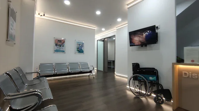 A hospital waiting room with metal chairs, a wheelchair, a flat-screen TV, and informational posters on the wall.