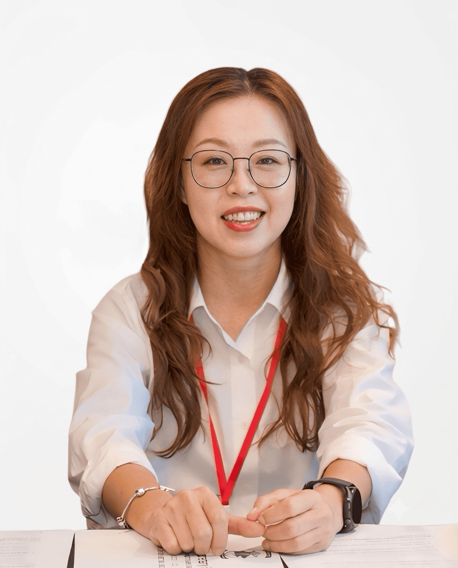 A woman with long red hair, wearing glasses, a white shirt, and a red lanyard, sitting at a desk with some papers.