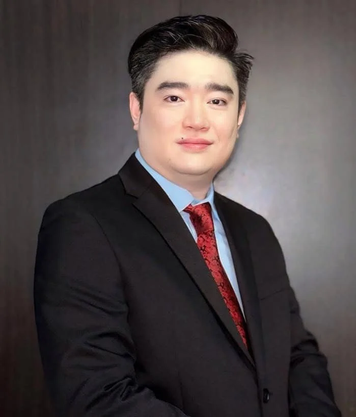 A portrait of a young man with dark hair wearing a black suit, light blue shirt, and red tie with a black pattern, standing in front of a dark background.
Dr. Khaing Lynn Maung, CEO of Permai Polyclinics Group
