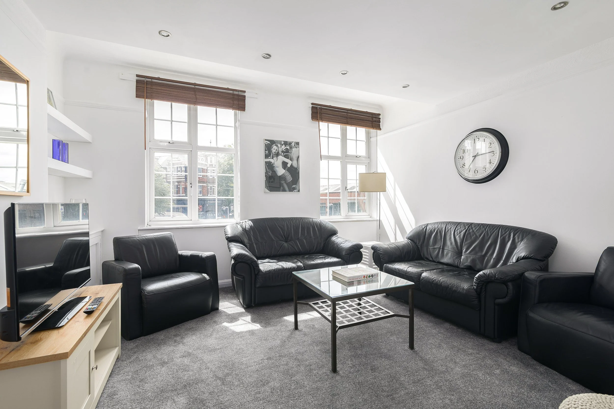A living room with white walls, large windows covered with brown blinds, black leather sofas, a glass coffee table, and a clock on the wall. There is a black and white photo hanging between the windows, and a TV with remote controls on a wooden stand