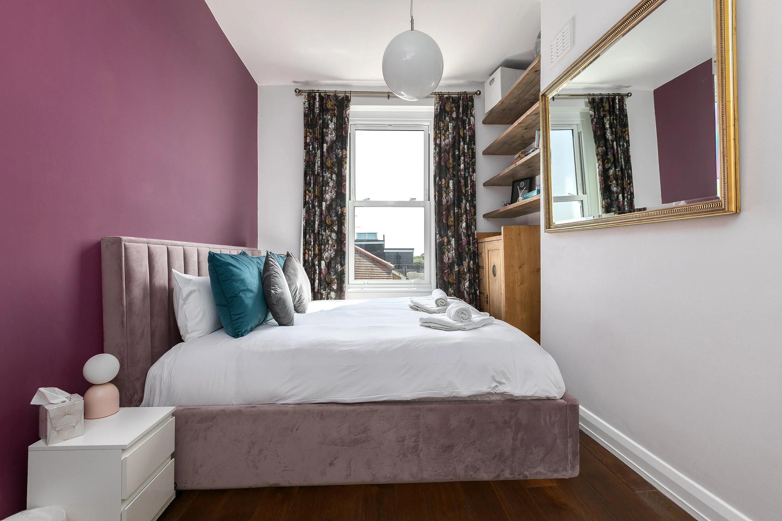A bedroom with a pink velvet bed frame, white bedding, and teal and gray pillows. A white nightstand with decorative objects, including a tissue box and a lamp. Two shelves with books and decorations above the window. A large mirror with a gold frame