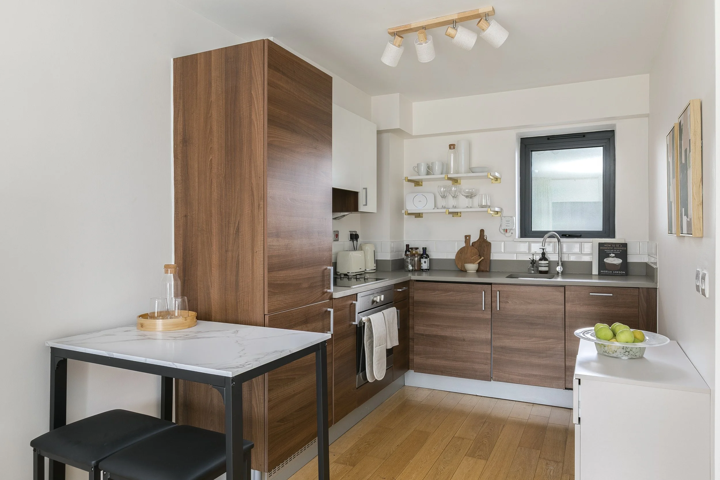 A kitchen with white walls, wooden cabinets, a small white table with black legs, and two black chairs. Open shelves with glassware and mugs are above the counter, which has a window behind it. A fruit bowl with green apples is on a white table in th