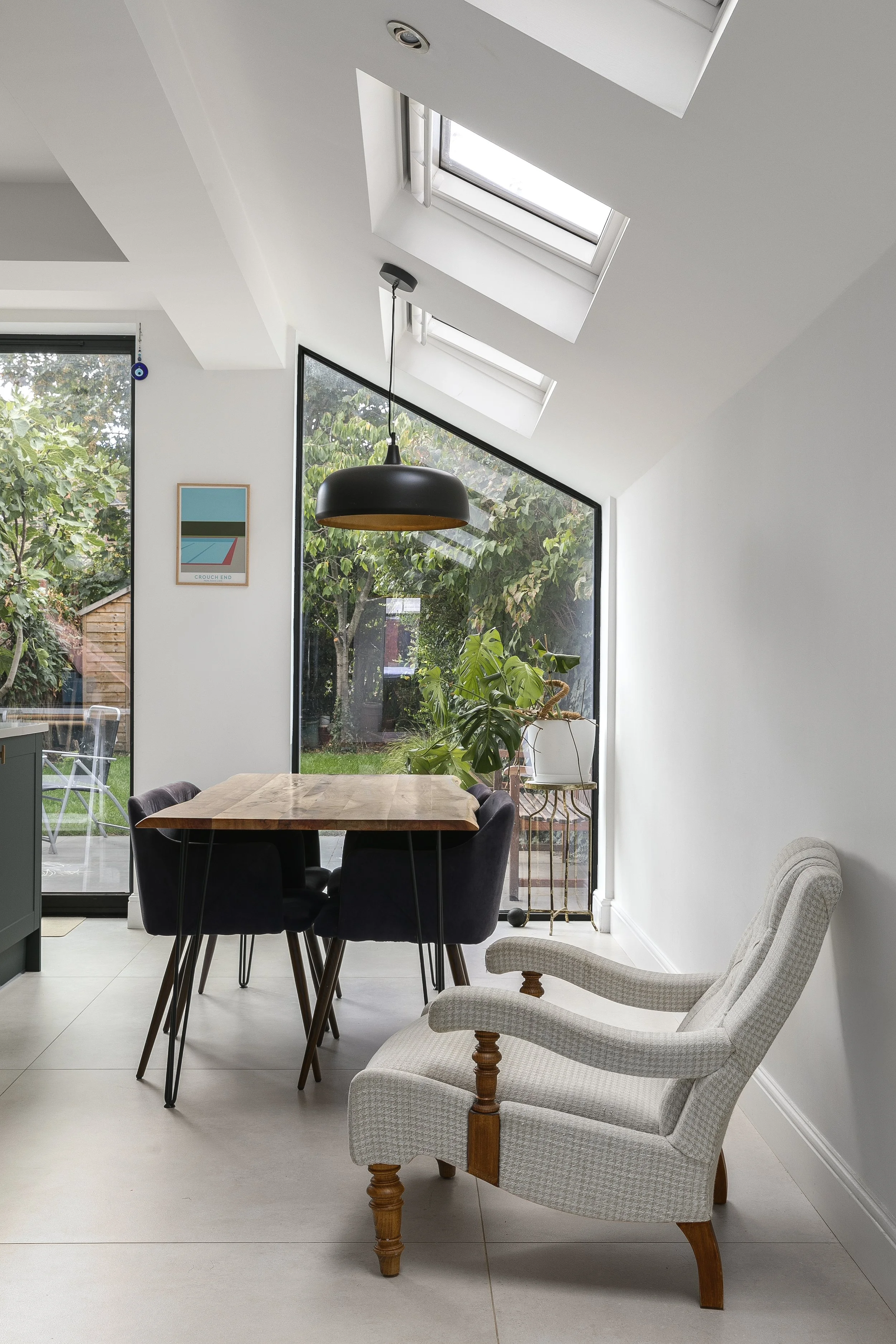 A modern dining room with a wooden table surrounded by five black upholstered chairs. The room has large windows and skylights allowing natural light, a decorative houseplant on a gold stand, and a framed art piece on the wall. There is a white, upho