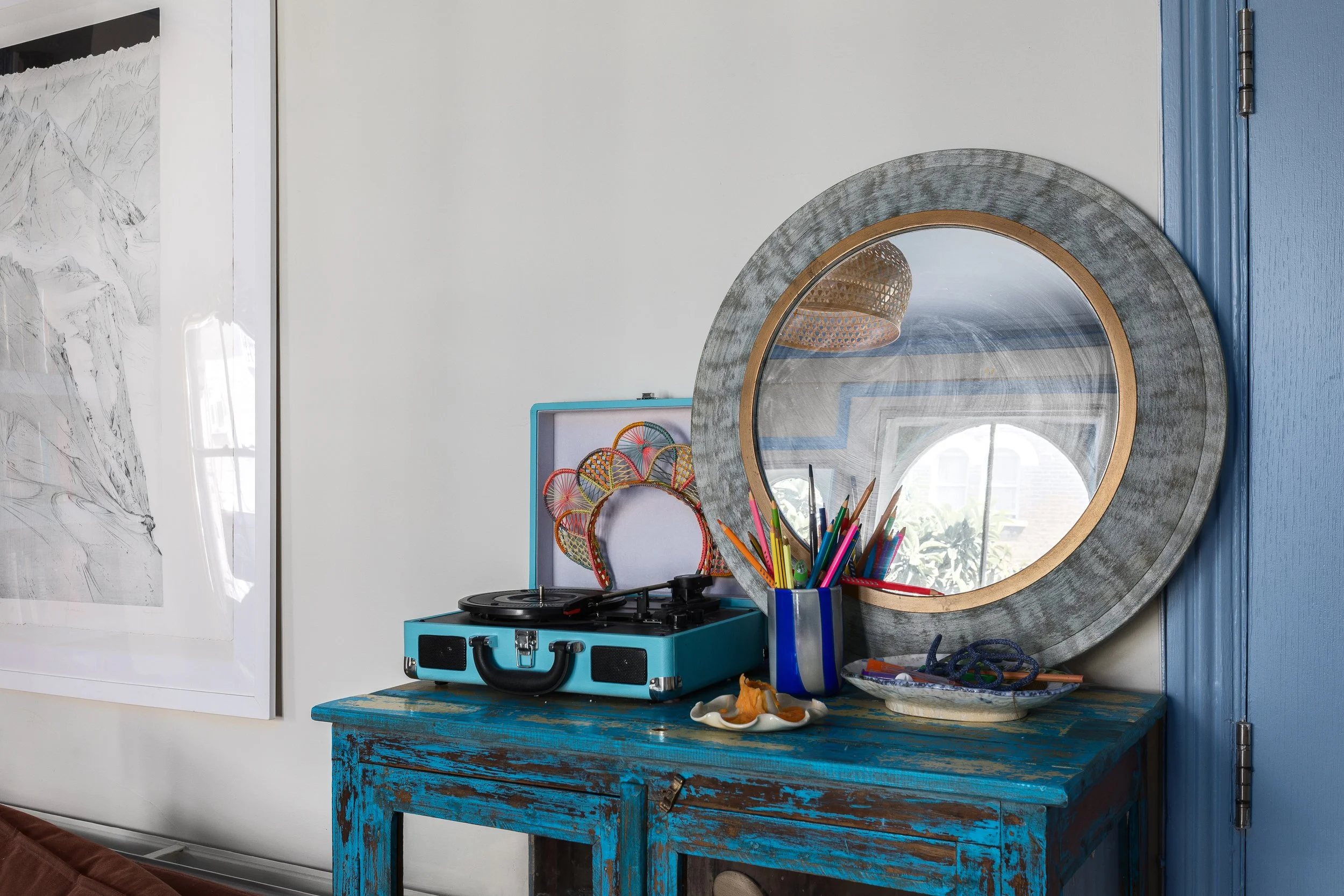 A round mirror with a gray and gold frame hangs on a wall above a distressed turquoise table. On the table, there is a vintage blue portable record player, a container filled with colorful pens and pencils, and a small white dish with orange items. P
