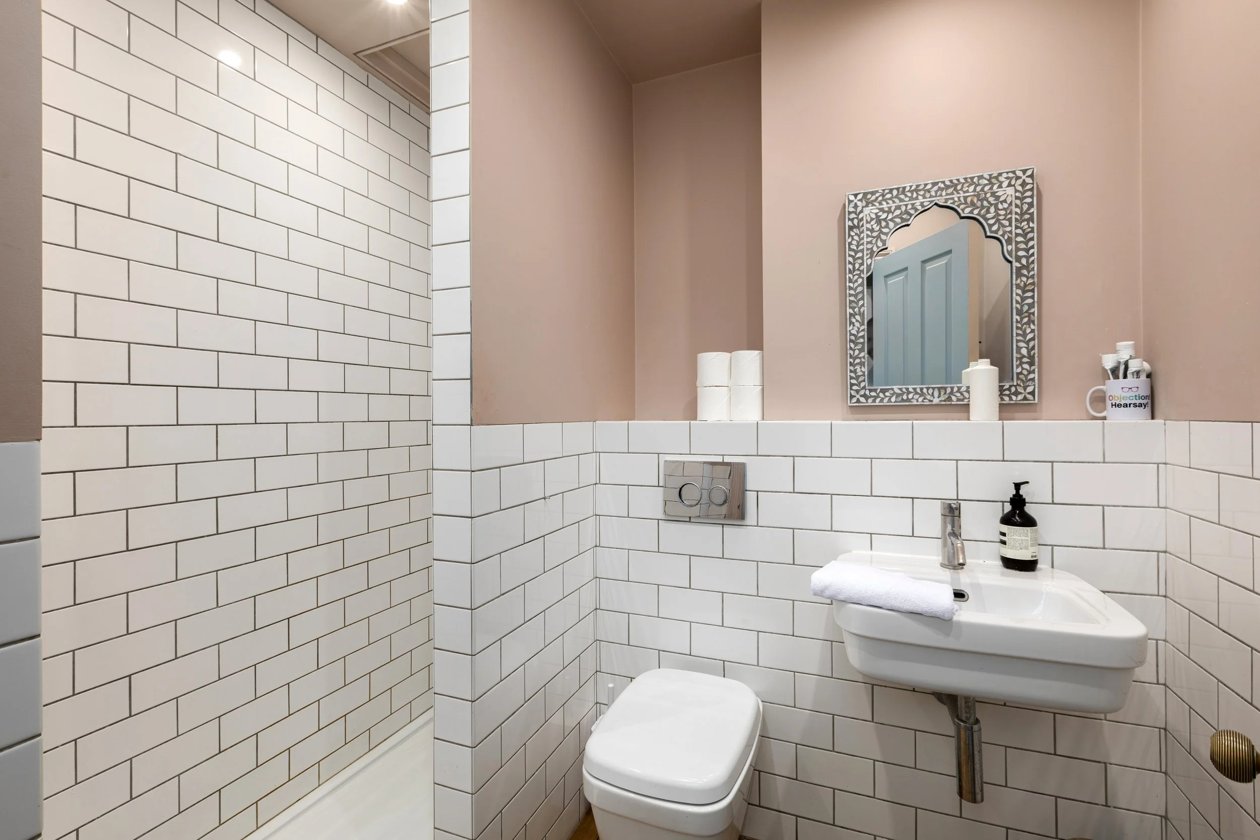 A small bathroom with white subway tile walls and a pink painted wall. There is a mirror with an intricate frame above a small white sink with a black soap dispenser. The bathroom has a toilet with a wall-mounted flush plate, and a shelf above the to