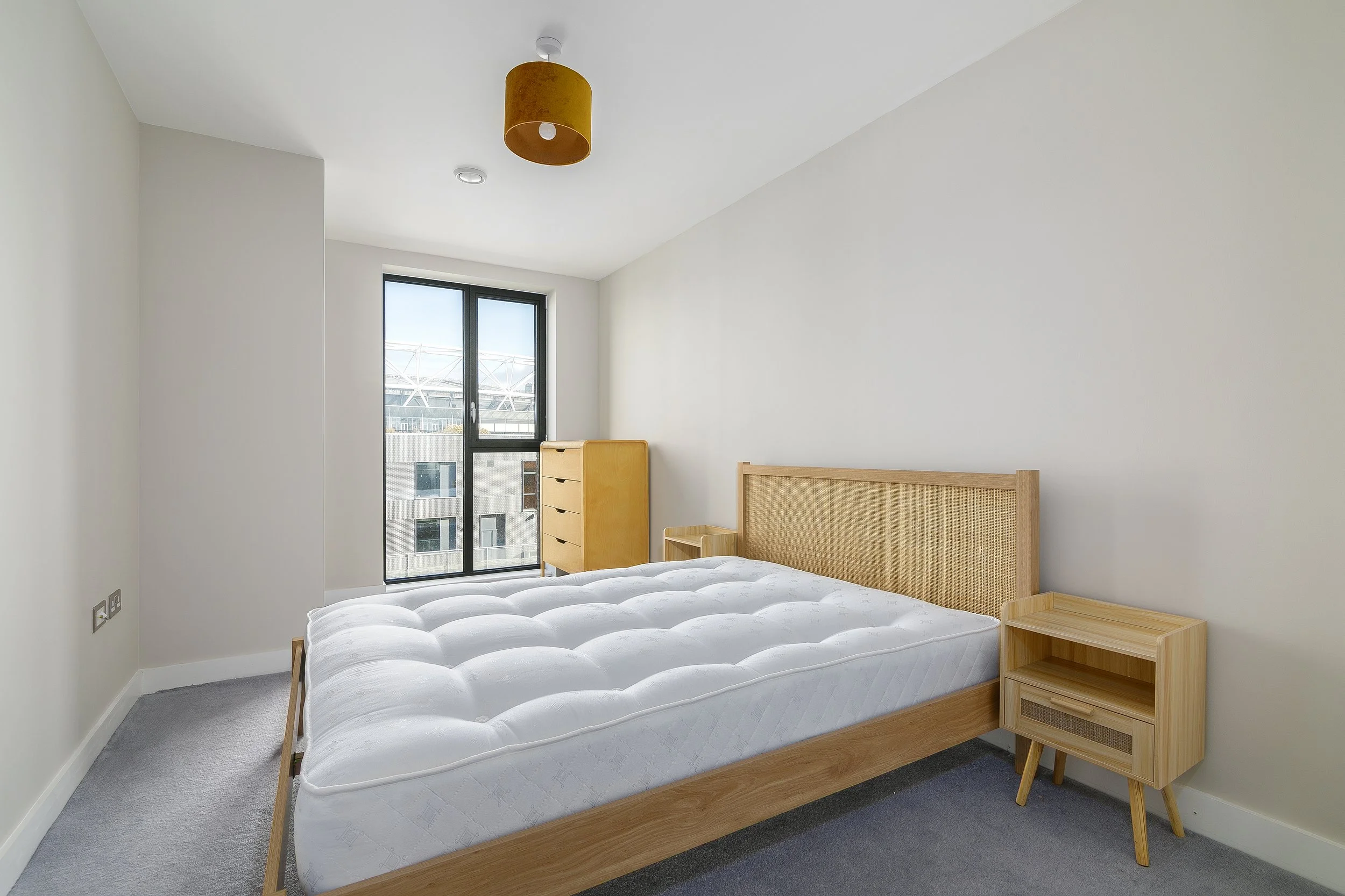 A minimalist bedroom with a white wall and a large window. There is a wooden bed frame with a white mattress, a small wooden bedside table, and a small wooden dresser. A mustard-colored lampshade hangs from the ceiling.