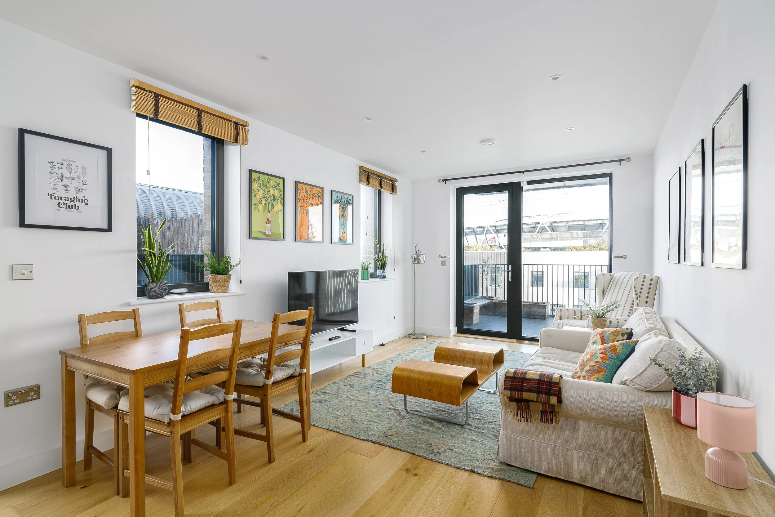 A bright living room with white walls, large windows, and wooden floors. It features a wooden dining table with chairs, a white TV stand with a flat-screen TV, a beige sofa with colorful cushions, artwork on the walls, houseplants by the windows, and