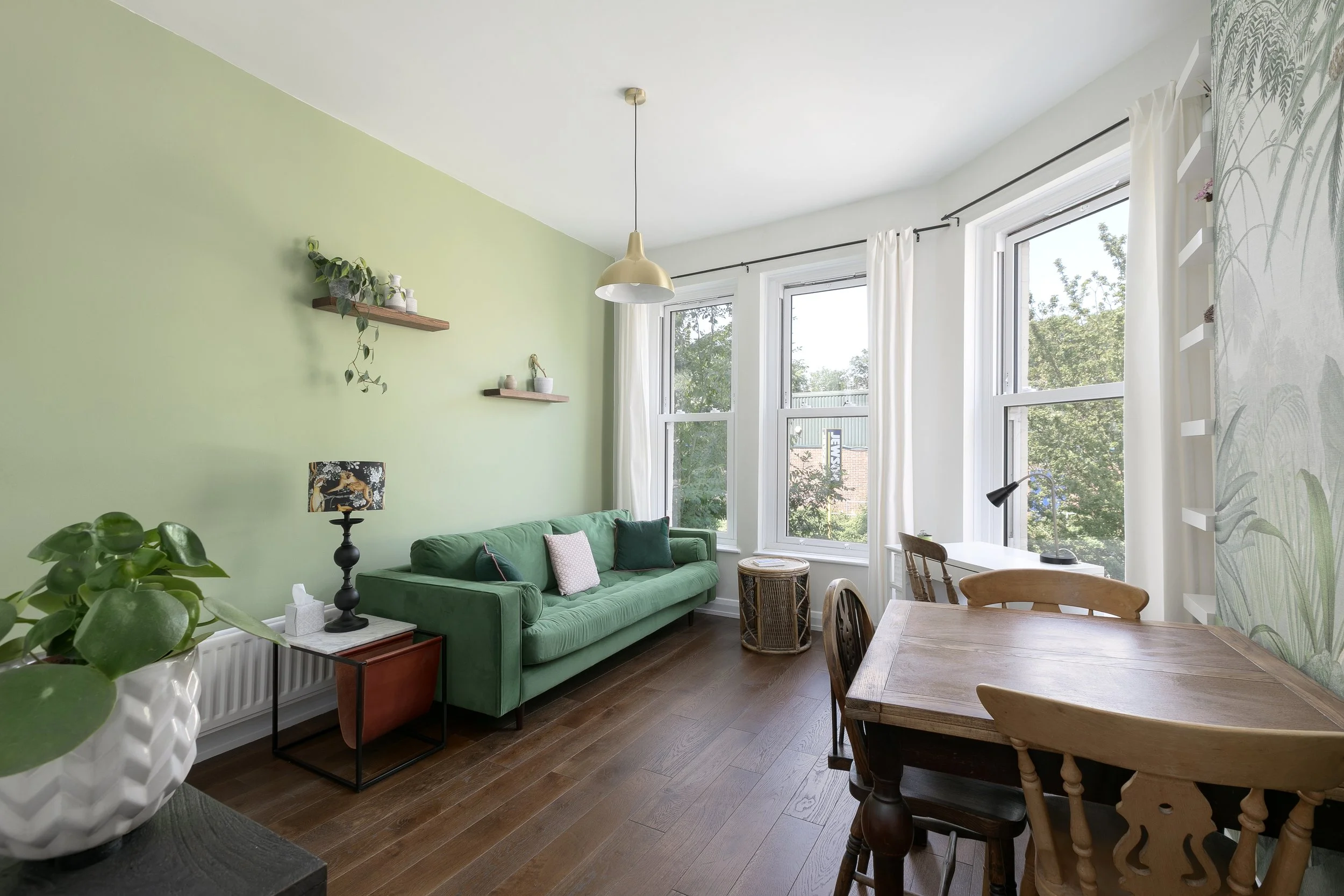 A living room with a green sofa, wooden dining table with matching chairs, large windows with white curtains, leafy plants, and a green accent wall with floating shelves and decorative items.