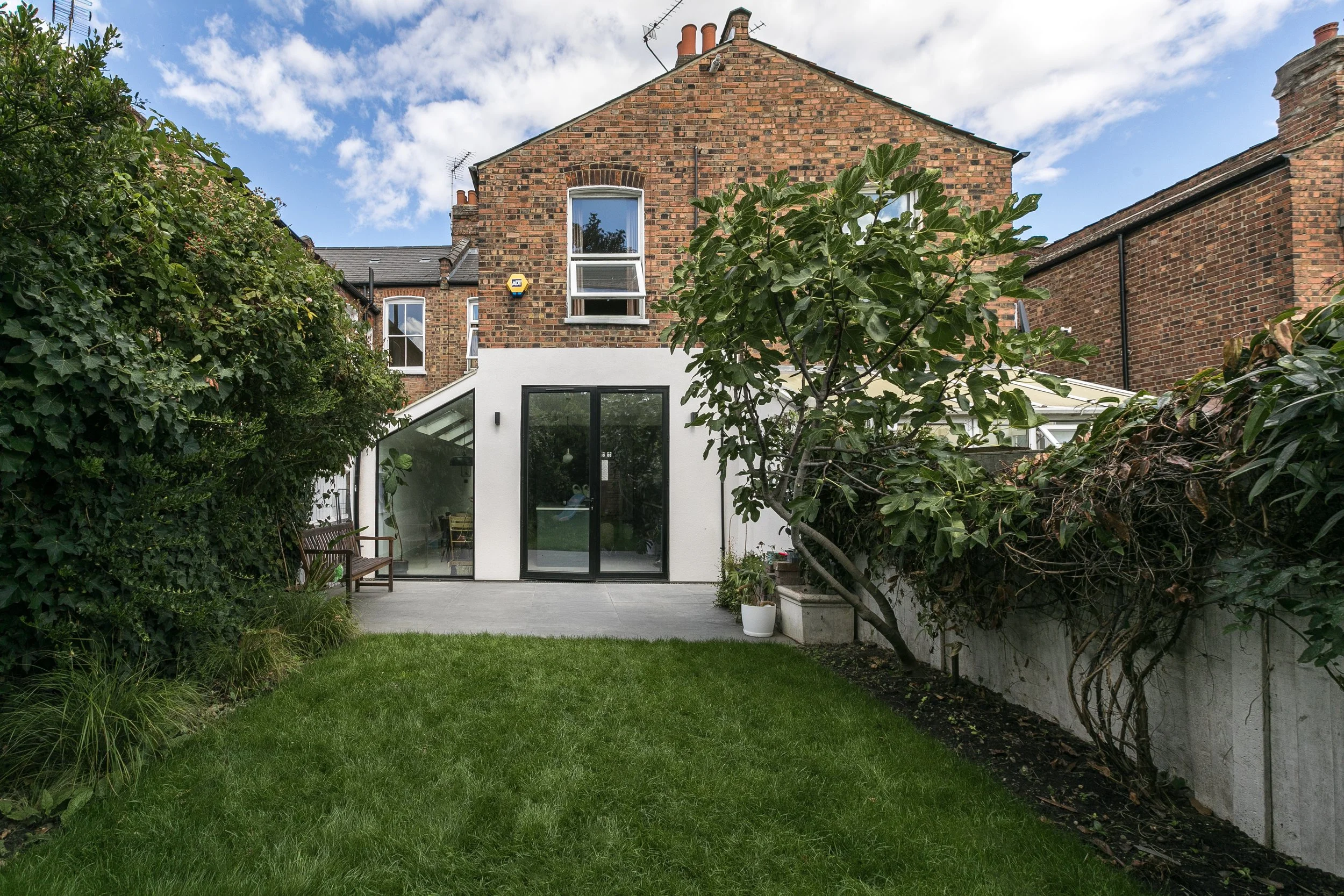 Backyard garden with lush green grass, tall leafy trees and shrubs, a white modern extension with large glass doors, brick houses with chimney stacks in the background, blue sky with clouds.