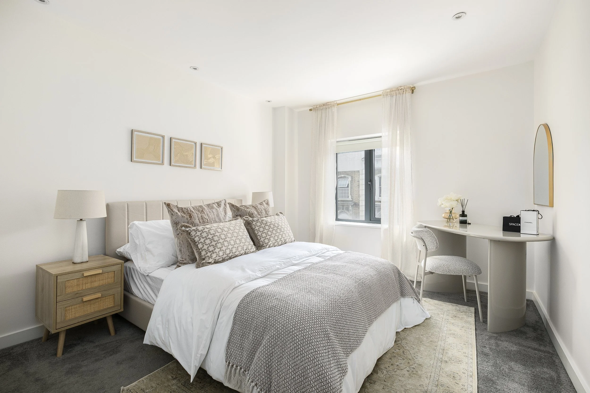 A minimalist bedroom with a white bed, beige decorative pillows, a wooden nightstand with a white lamp, and a small desk with a chair near a window with sheer curtains. The room has light-colored walls, framed artwork, and a mirror on the wall.