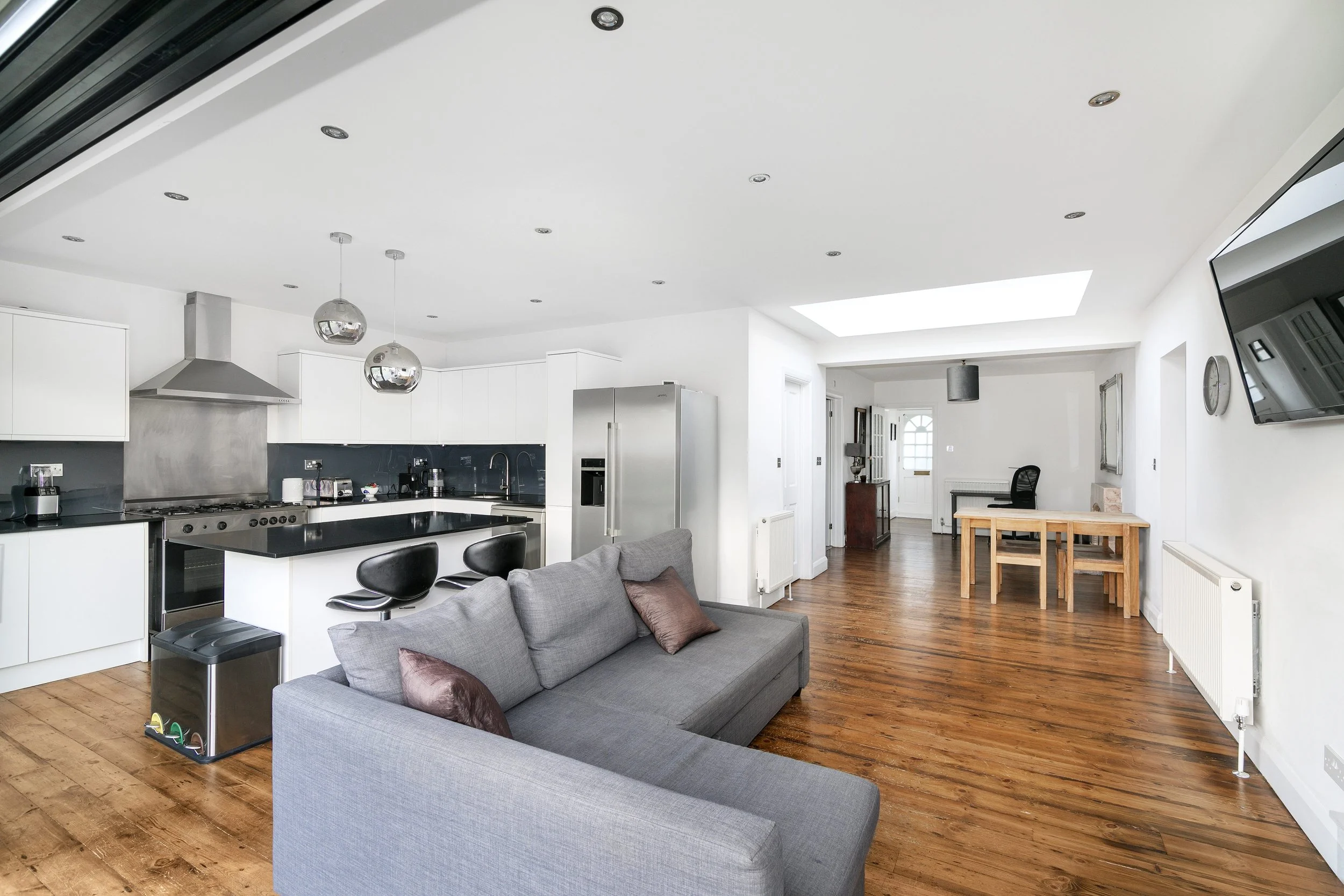 Modern open-concept living room and kitchen with white walls, wooden flooring, gray sofa, black kitchen island, white cabinets, stainless steel appliances, pendant lights, and a wall-mounted TV.