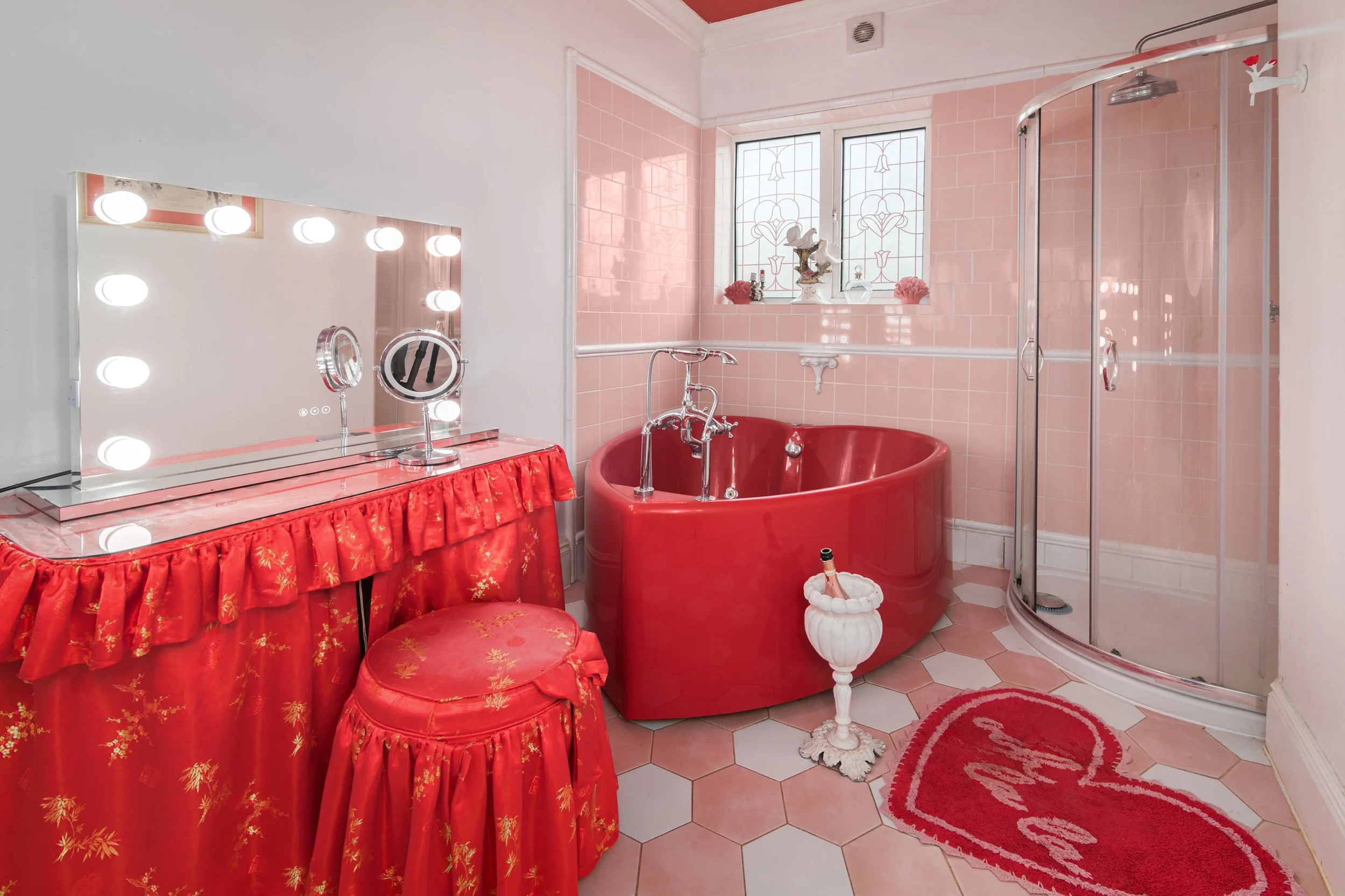 A bathroom decorated in pink with a red bathtub, pink tiled walls, and a pink and red rug. There is a vanity mirror with lights, a red chair with a matching ottoman, and a glass-enclosed shower with curved doors. The window has stained glass and deco