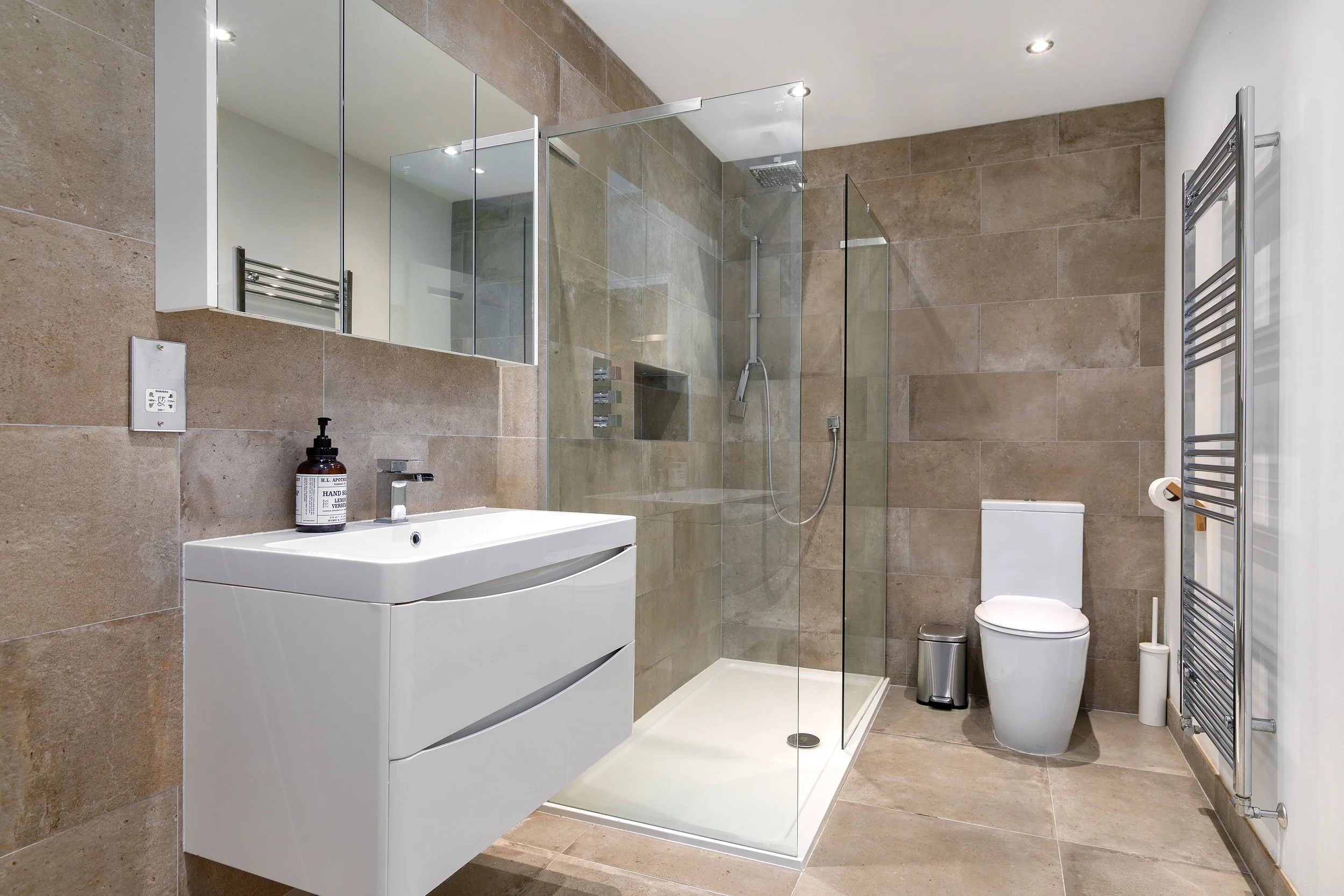 Modern bathroom with beige tiled walls and floor, featuring a walk-in shower with glass panels, a white wall-mounted sink, a mirror, a toilet, a small trash can, a towel radiator, and a source of natural light.