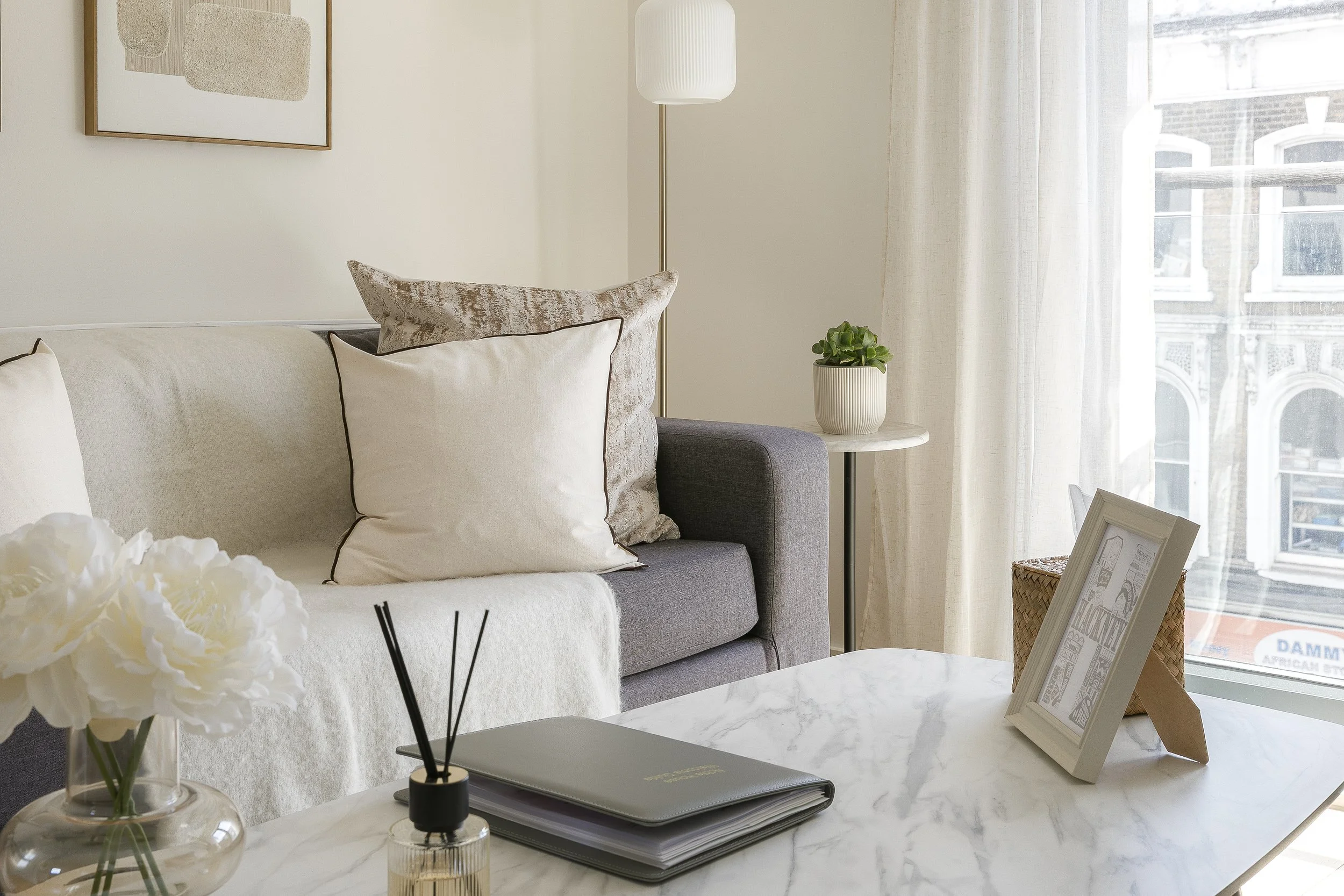 A living room corner with a light gray sofa adorned with beige and patterned throw pillows, next to a white side table with a potted plant. A floor lamp with a white shade stands beside the sofa. Light filtering through sheer curtains reveals a view 