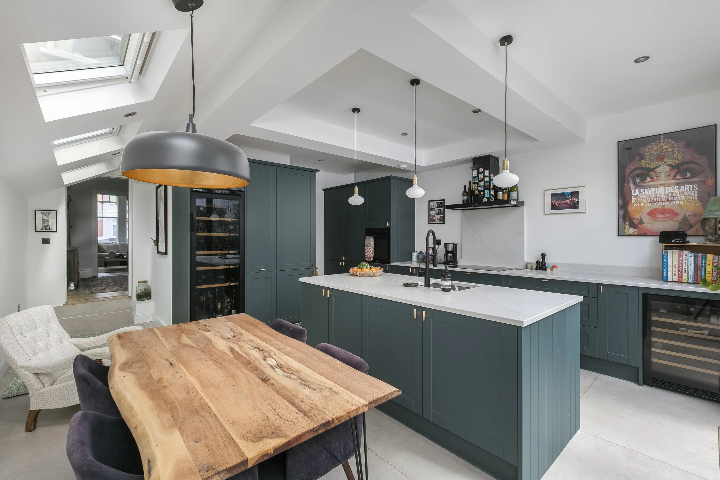 A modern kitchen with a white marble island, teal cabinets, and pendant lights. There is a dining table with a wooden top and chairs, and skylights providing natural light.