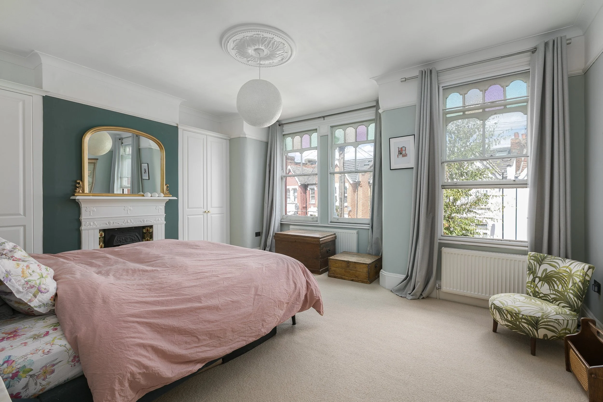 A bedroom with a pink bedspread, floral sheets, a white fireplace with a gold-framed mirror, large bay windows with stained glass details, and light-colored curtains. There are wooden trunks and a patterned chair near the windows.