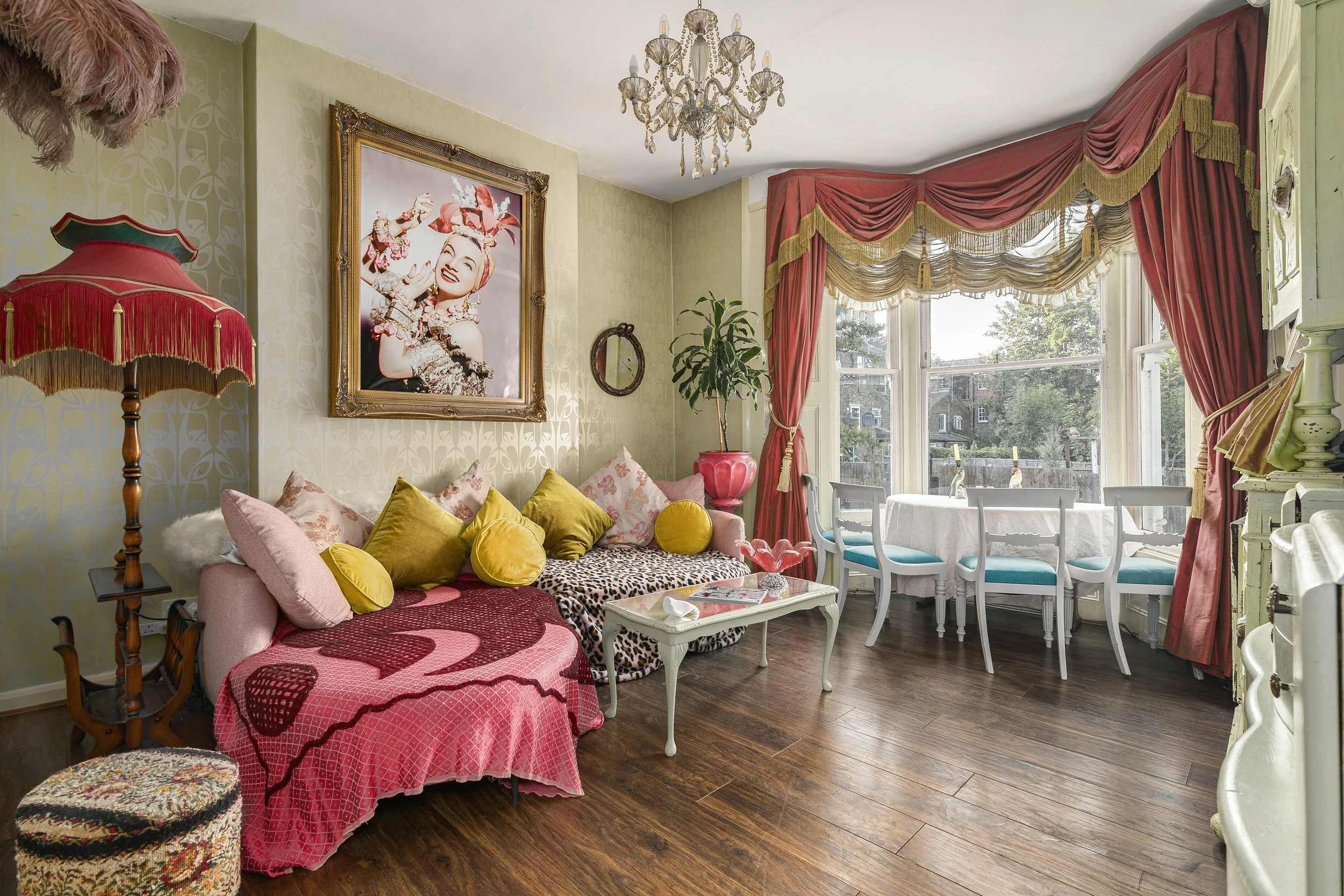 A cozy vintage living room with a pink sofa covered in yellow and pink pillows, a leopard print blanket, and a small footstool. There's a large framed picture of a woman with a red headscarf, ornate chandelier, potted plant, and tall window with red 