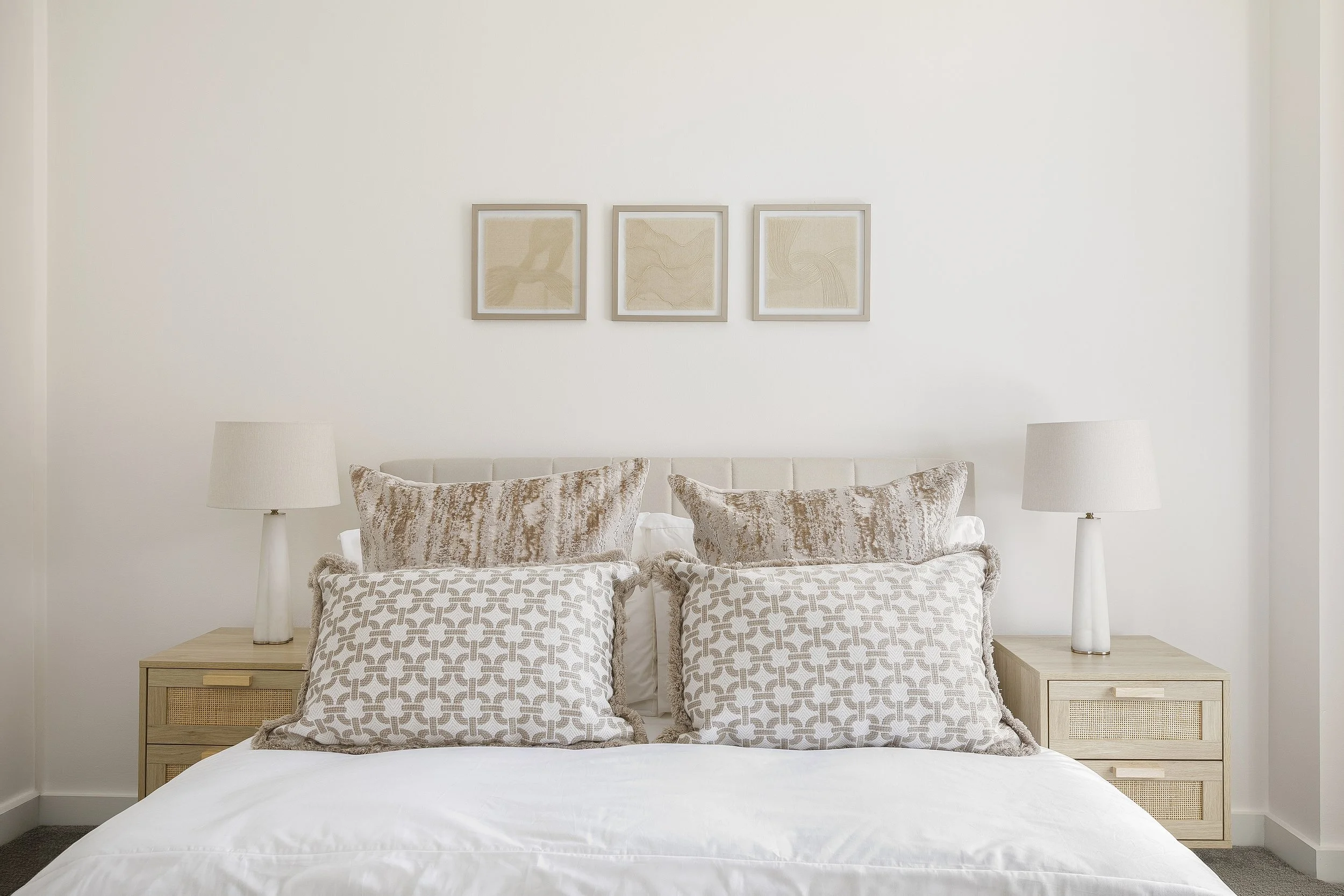A neatly made bed with beige patterned pillows, flanked by two identical white table lamps on wooden nightstands, set against a white wall with three framed abstract art pieces.