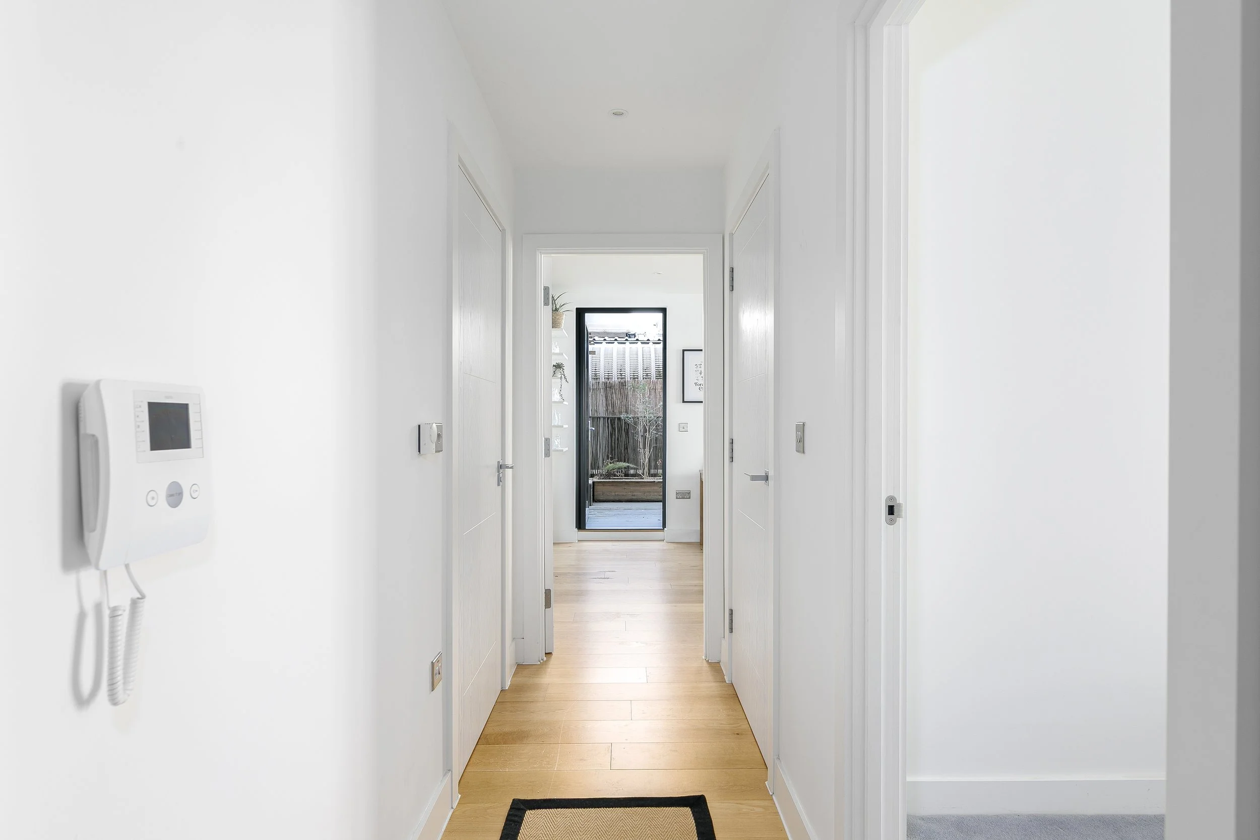 A bright white hallway with wooden flooring leading to a glass door outside, with a small black and beige rug at the entrance.