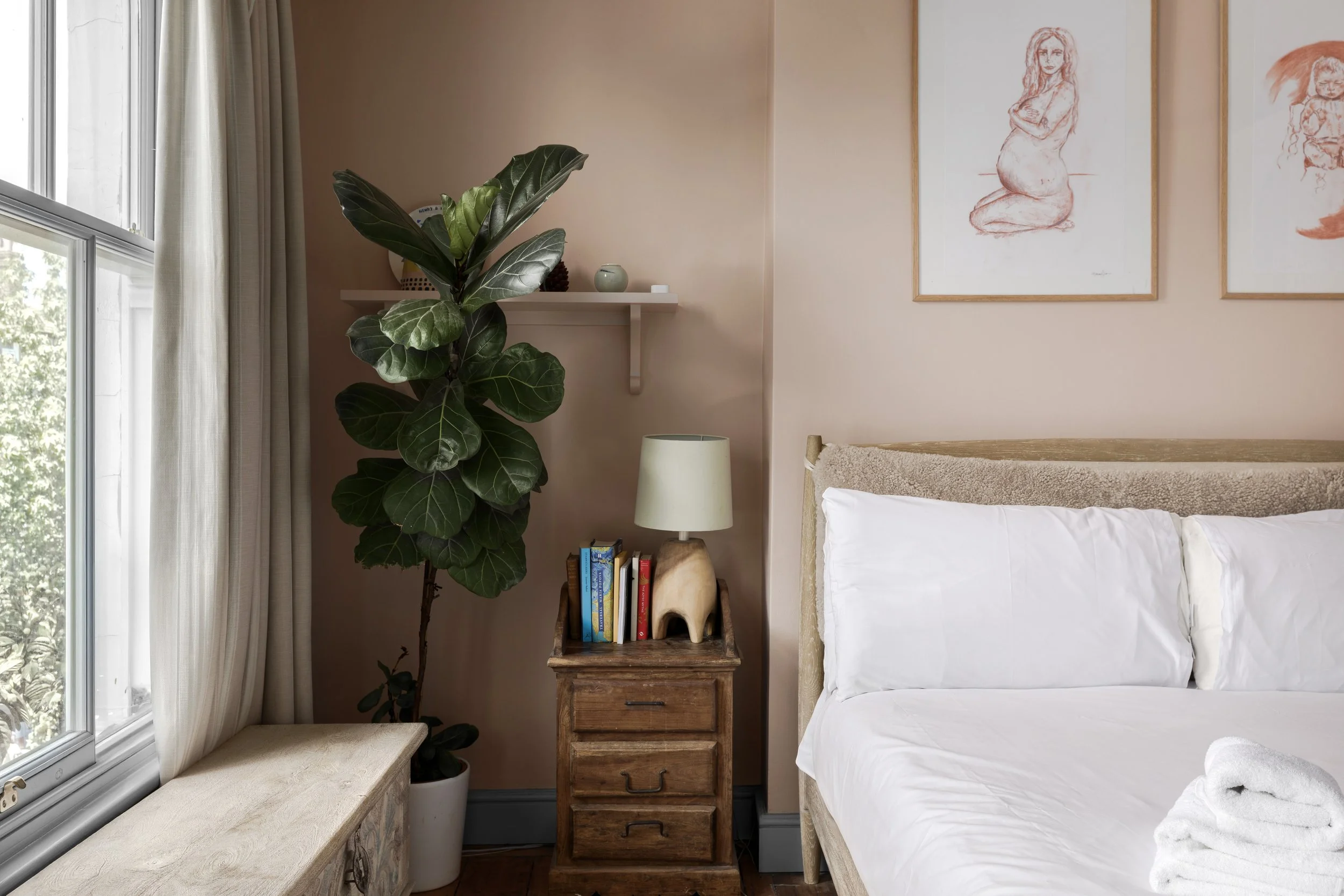 A cozy bedroom corner with a large potted fiddle leaf fig plant next to a window with off-white curtains. A white shelf with decorative items is mounted on the wall above a rustic wooden nightstand, paired with a beige bedside lamp. The bed has white linens, and above it are framed sketches of women in a minimalist style.