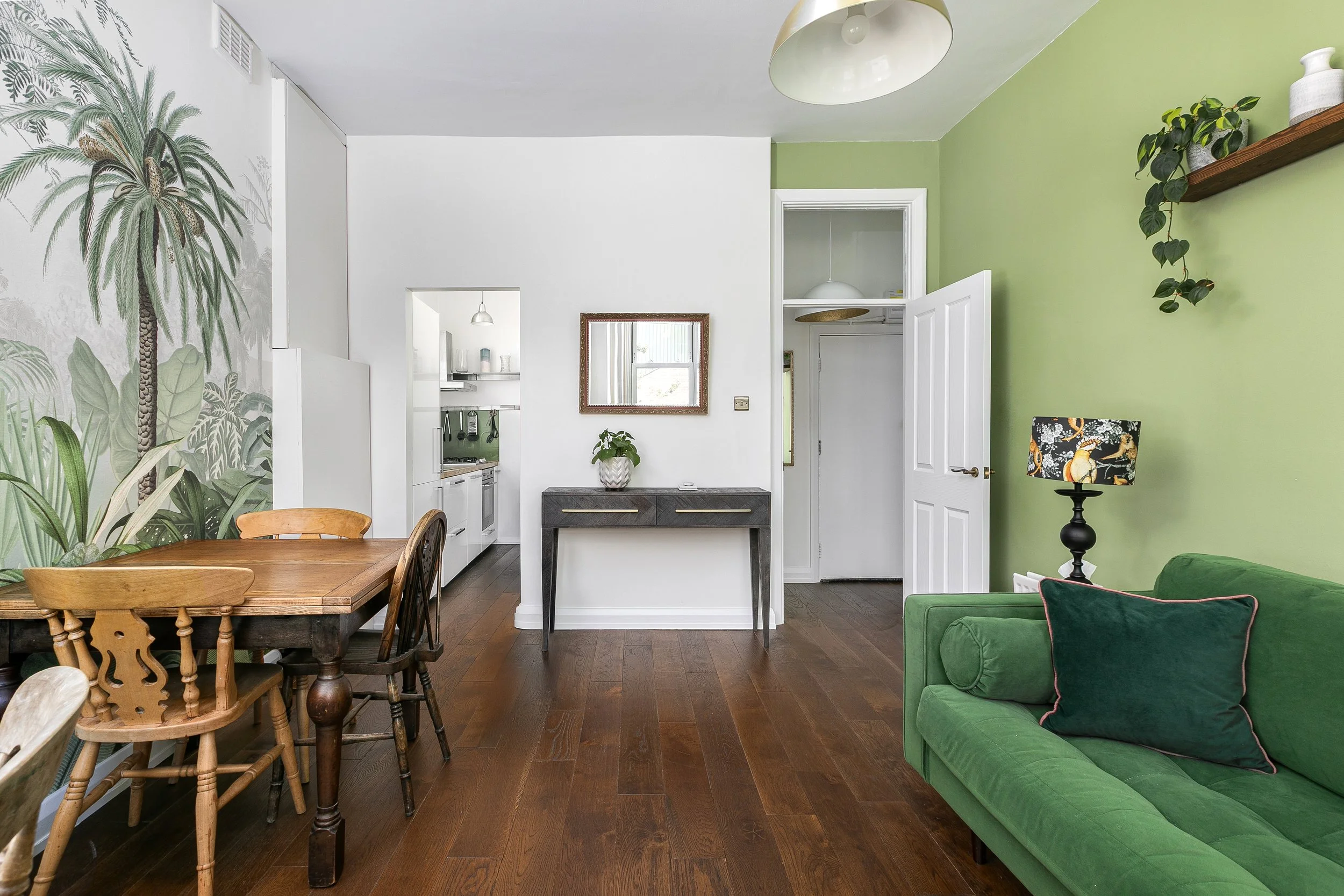 A living room with a green sofa, a wooden dining table with chairs, a floor lamp, a mirror, and a wall with green and white paint and decorative plants.